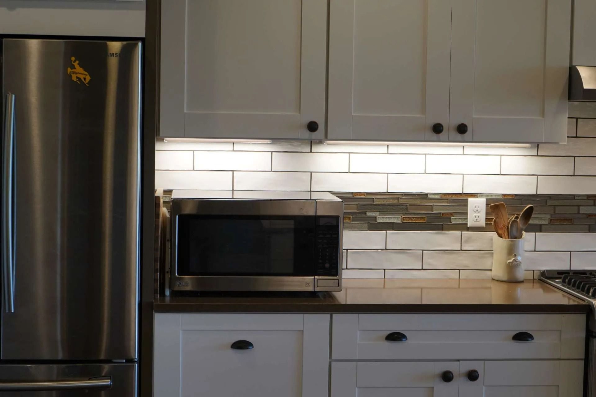 A stainless steel microwave sits on a brown countertop under white cabinets in a kitchen with a tiled backsplash.