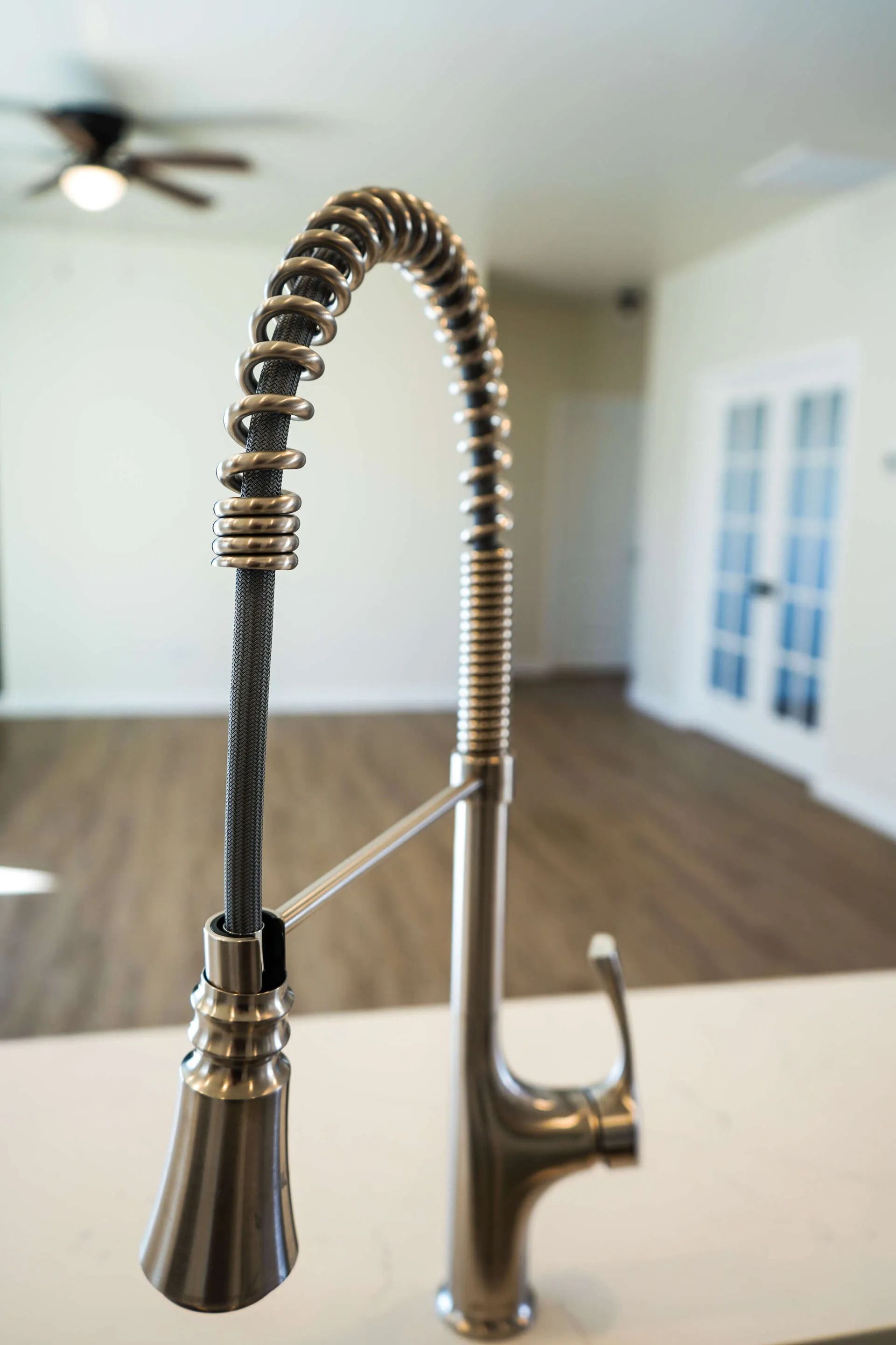 A brushed nickel kitchen faucet with a pull-down sprayer nozzle and coiled neck, set against a blurred room interior.