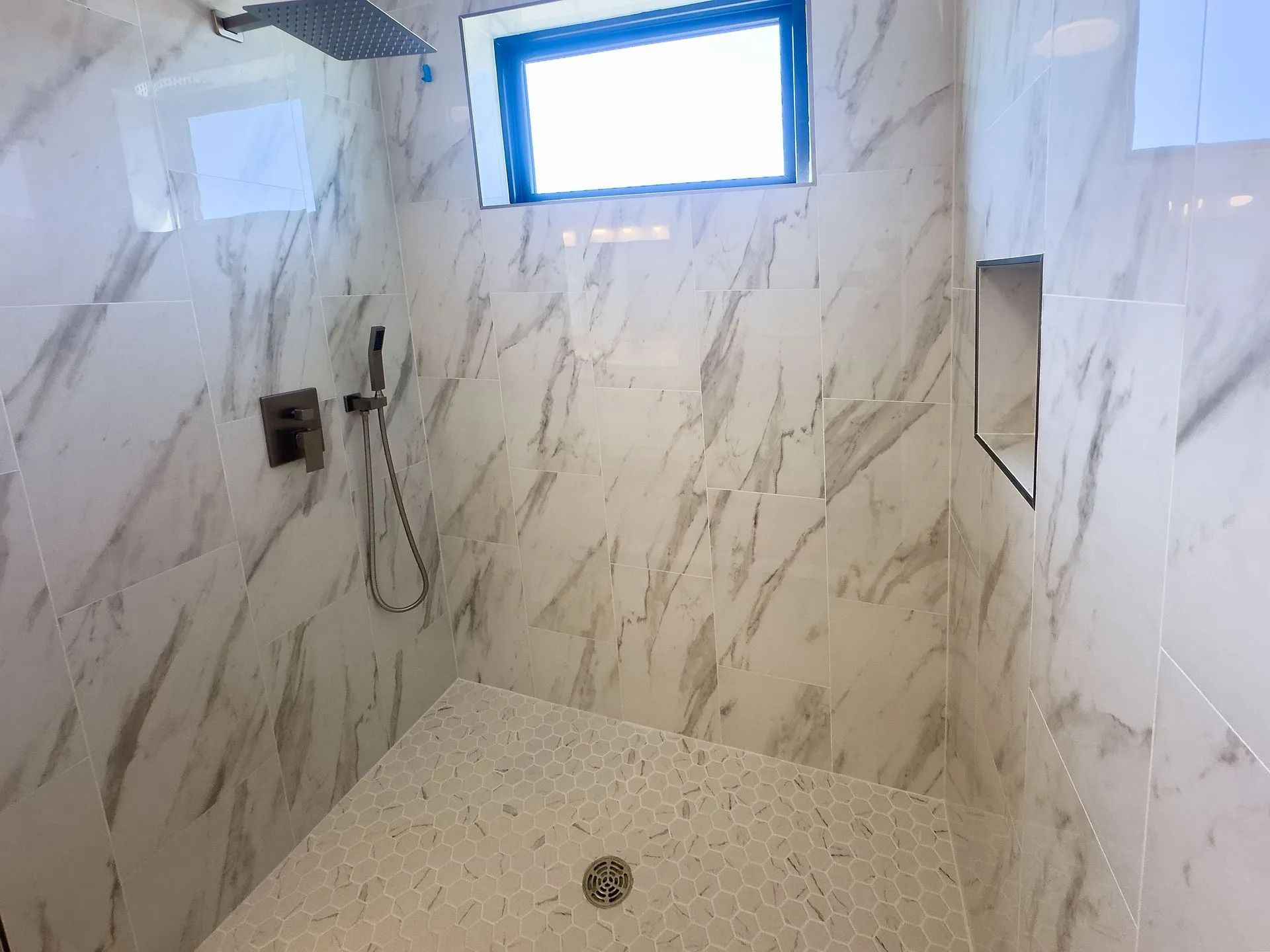 A modern walk-in shower with white marble-patterned tiled walls, a rain showerhead, a handheld sprayer, and a small window.