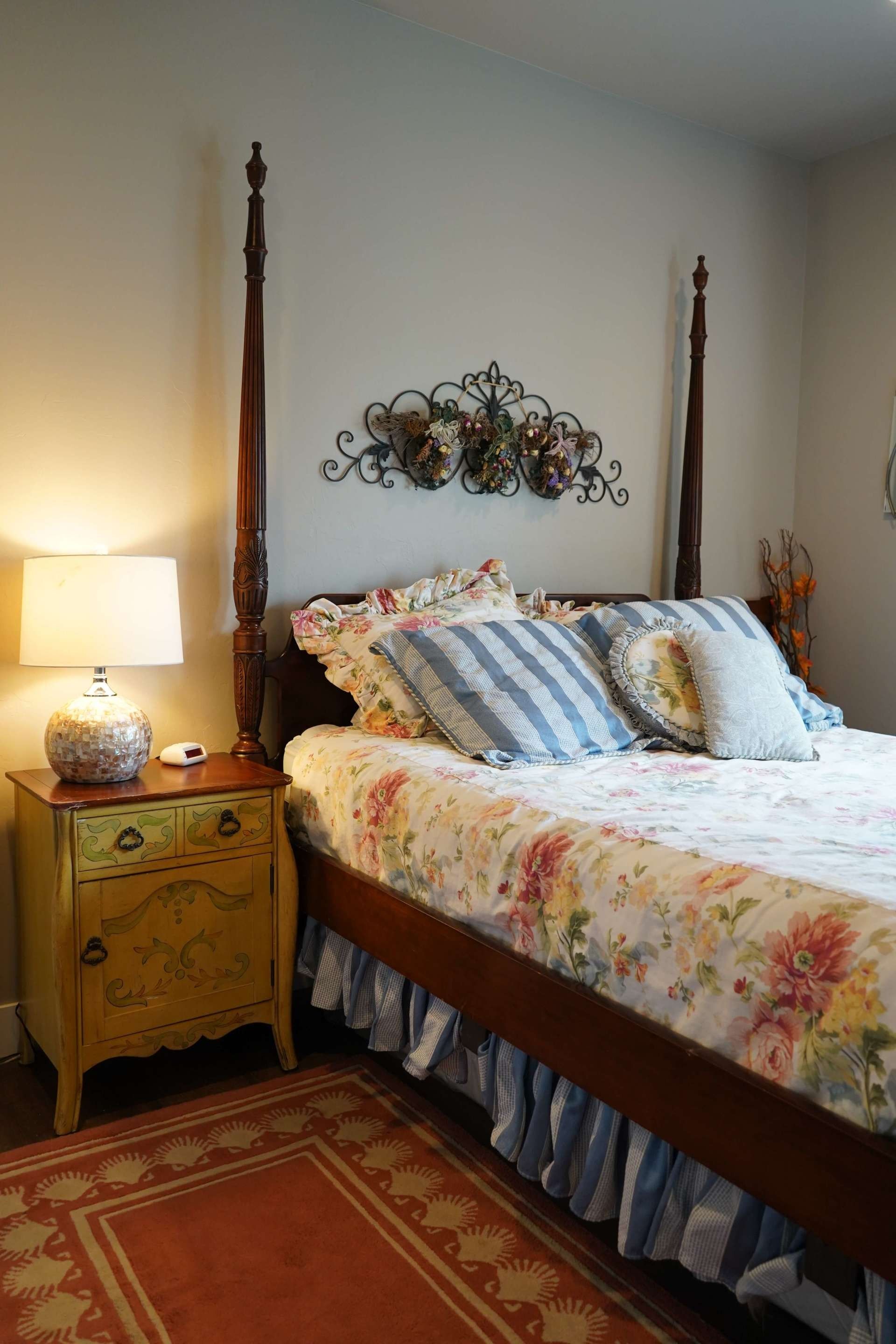A wooden four-poster bed with floral bedding and blue striped pillows sits next to a yellow nightstand with a lamp.