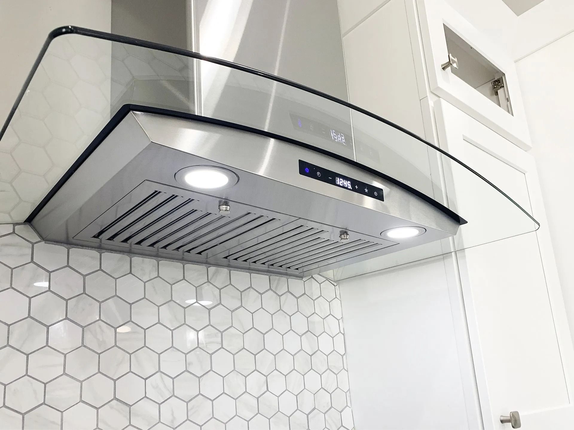 Stainless steel kitchen range hood with curved glass accents installed over a white hexagonal tile backsplash.