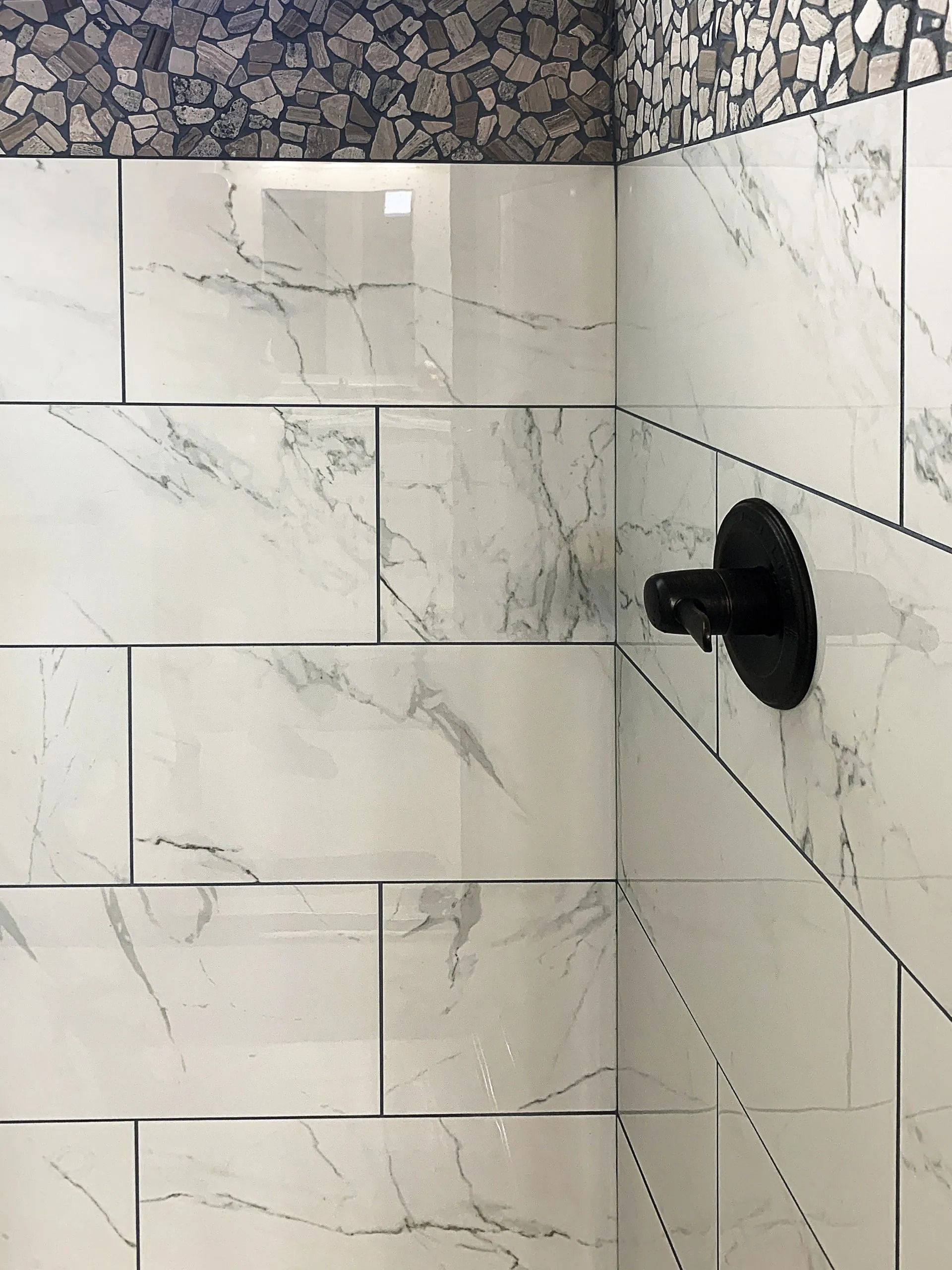White marble-look subway shower tiles with a dark stone-textured accent border and a black round shower control handle.