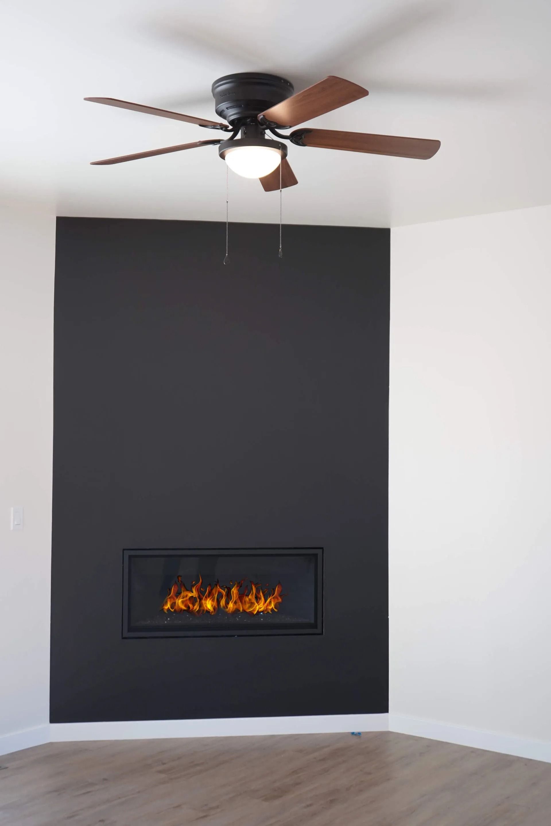 A modern corner fireplace with a black accent wall and a dark wood and black ceiling fan centered above in a room.