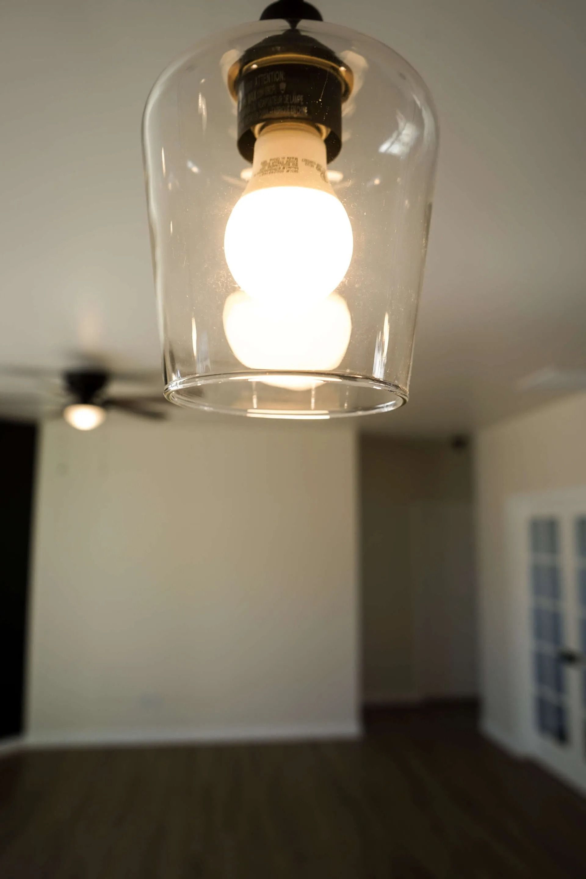 A lit glass pendant lamp hangs in the foreground with a blurred room featuring a ceiling fan and french doors behind it.