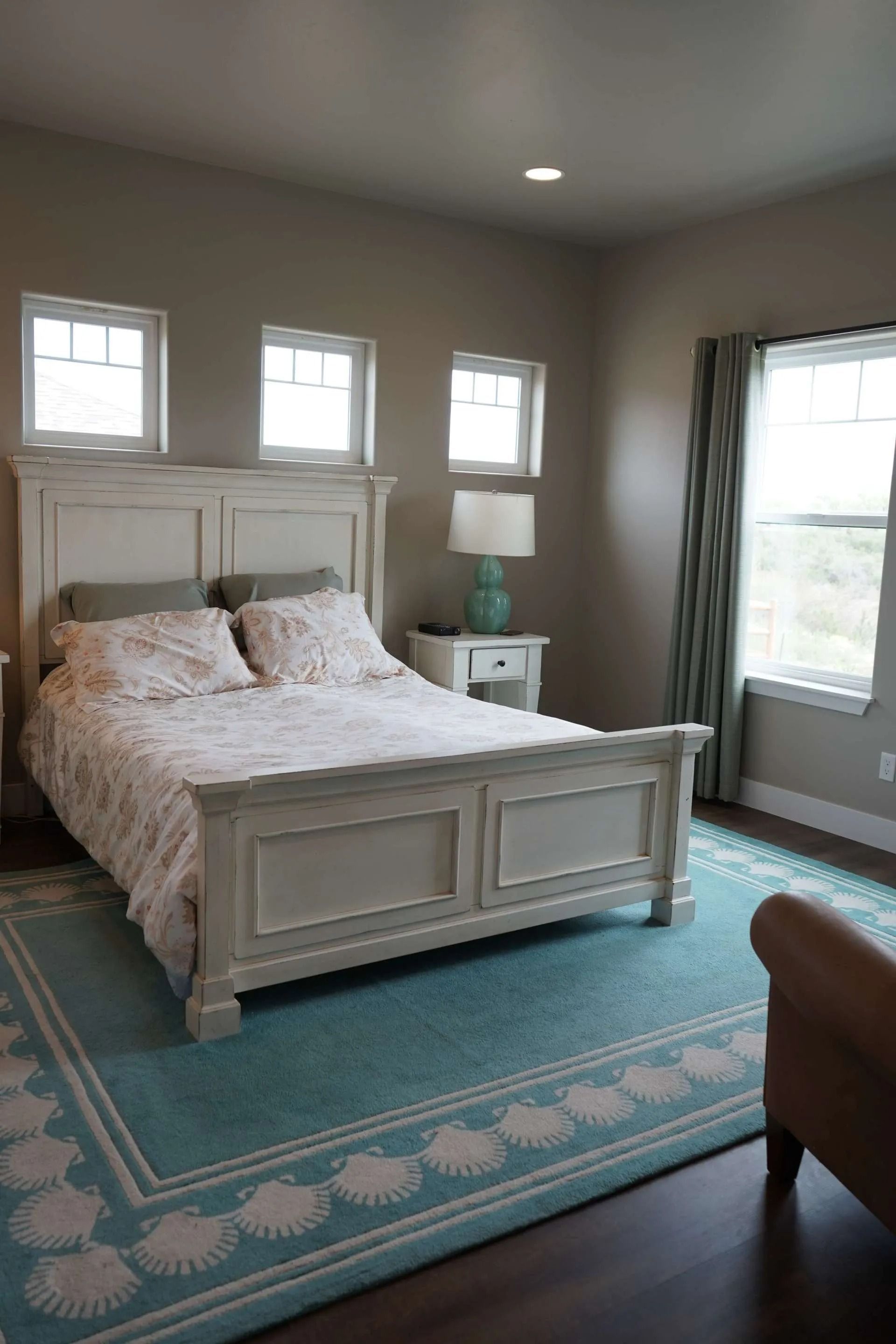 A bedroom with a white wooden bed, floral bedding, a turquoise patterned area rug, a bedside lamp, and window curtains.