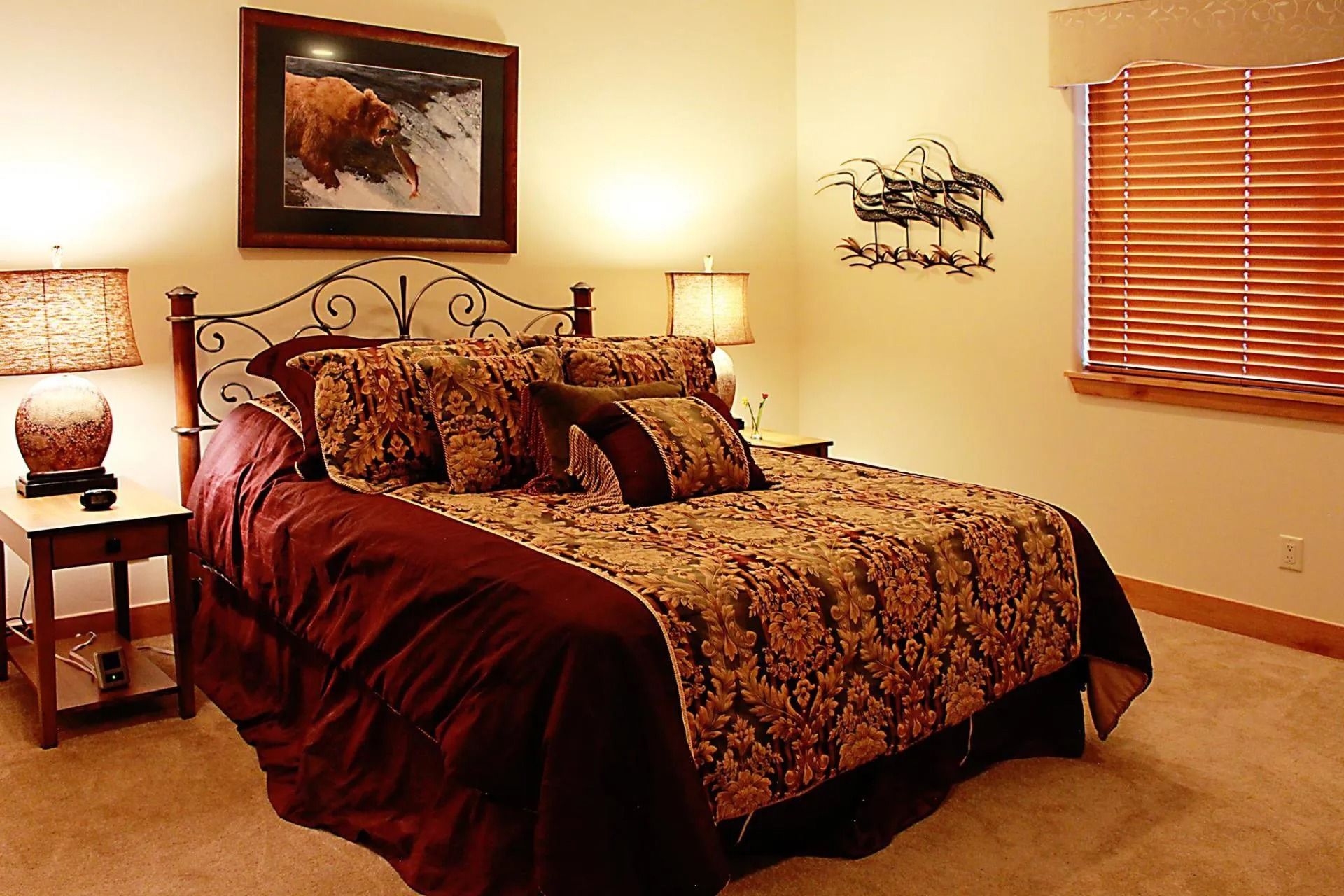 A bedroom features a queen bed with maroon and gold patterned bedding, a nightstand with a lamp, and wall art.