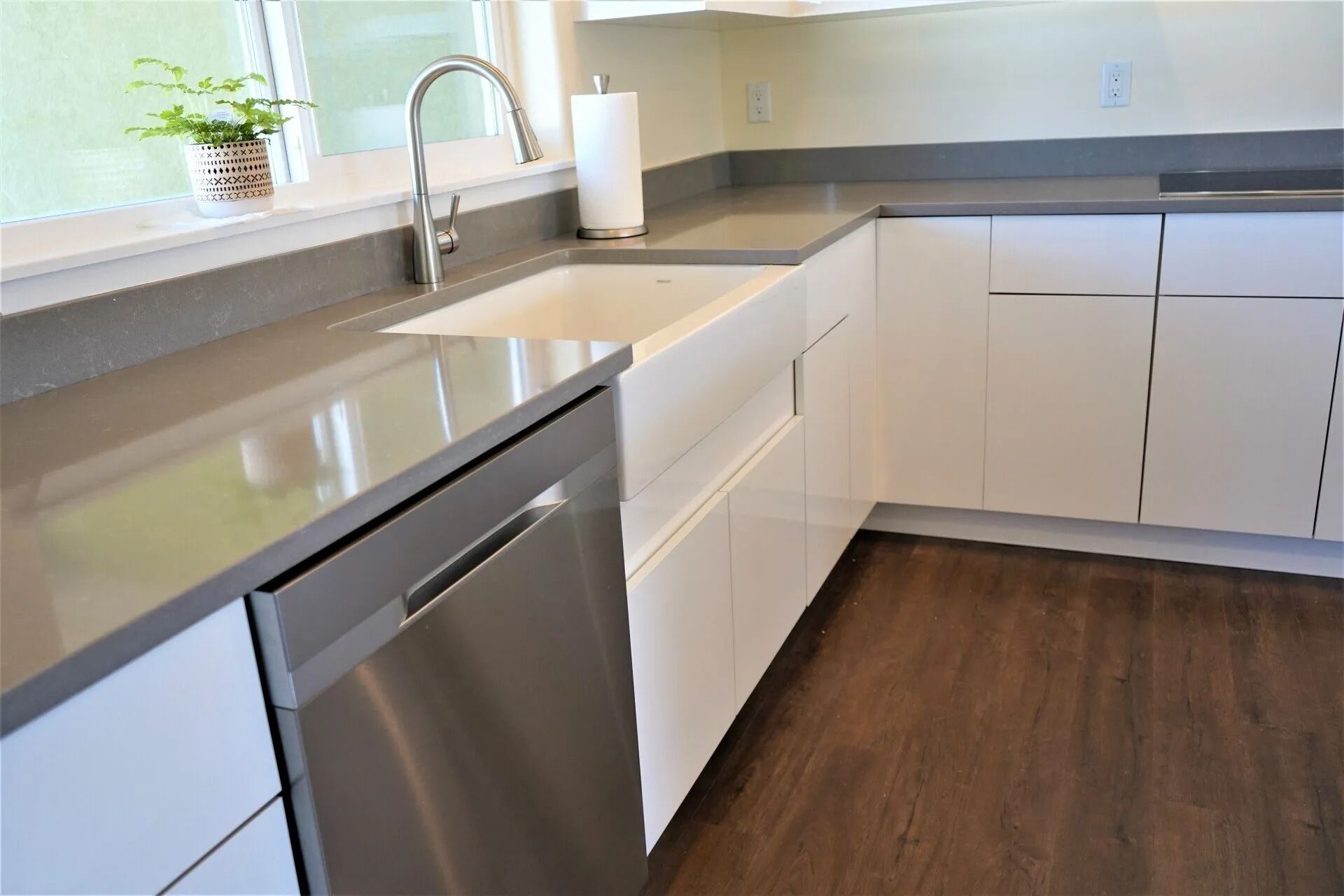 A modern kitchen with white cabinets, a farmhouse sink, stainless steel dishwasher, and grey countertops.
