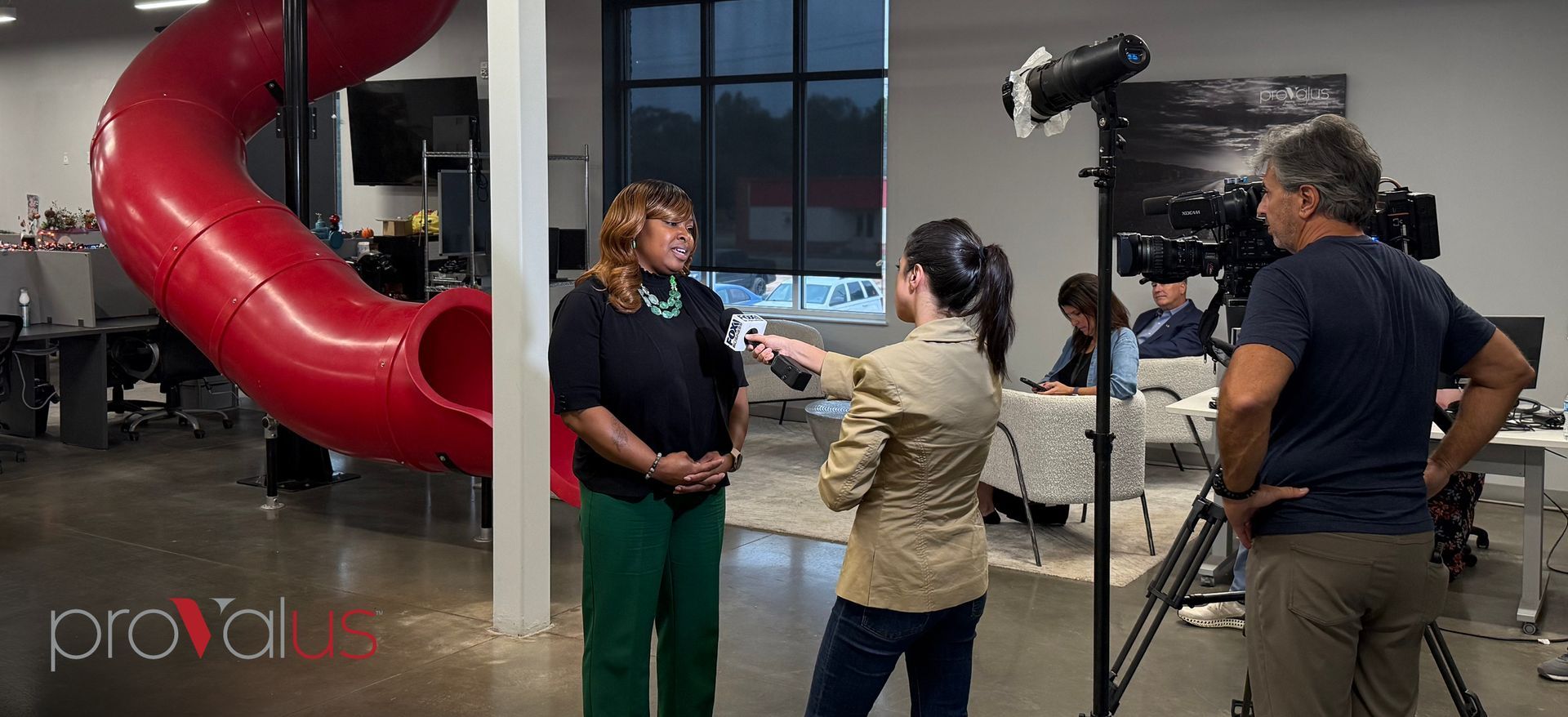 Two people interviewed in a studio beside a red spiral slide, with cameras and a tripod filming.