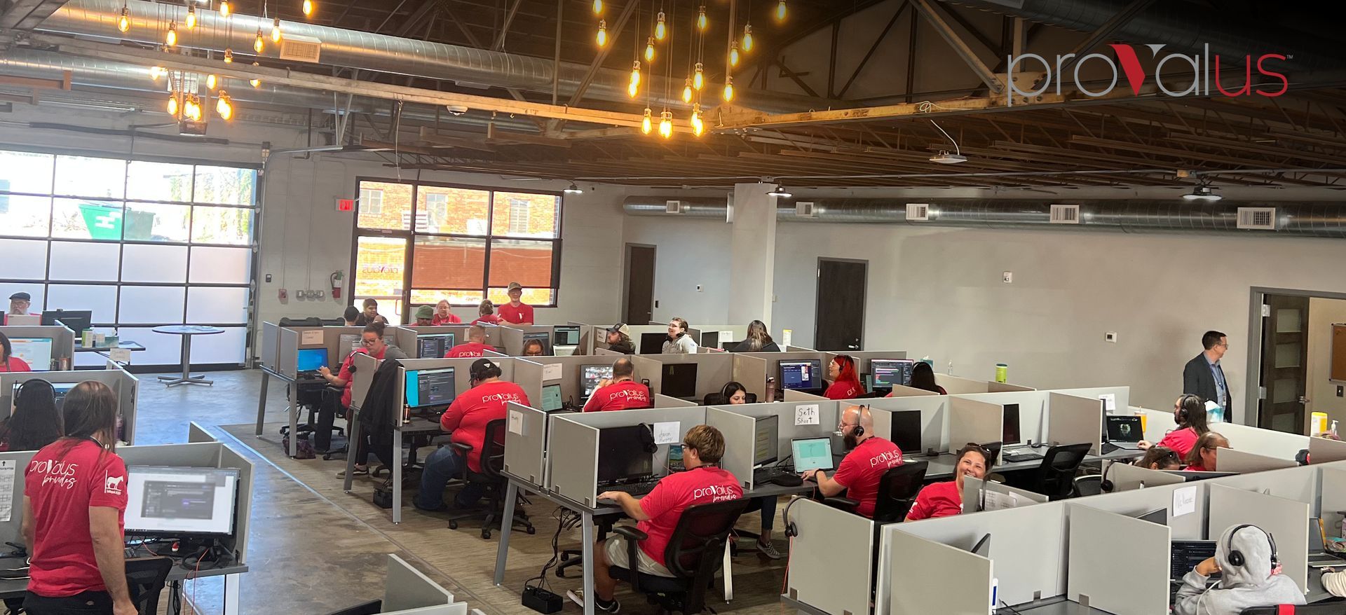 Open-plan office with employees in red shirts working at white desks under warm overhead lights