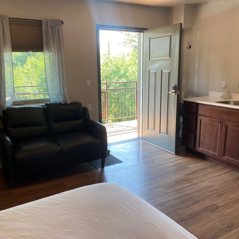 Small room with a black couch, open door to a balcony, and a small kitchenette.