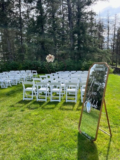 Wedding ceremony setup: white chairs on green grass with sign, tall trees in background.