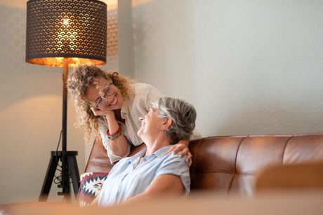 A woman with curly hair smiles while resting her chin on her hand behind another woman seated on a brown leather couch. A woman with curly hair smiles while resting her chin on her hand behind another woman seated on a brown leather couch.
