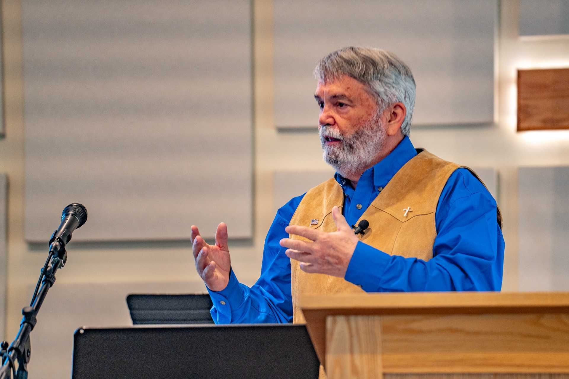Man with gray beard giving a presentation at a podium, gesturing with his hands. He's wearing a blue shirt and tan vest.
