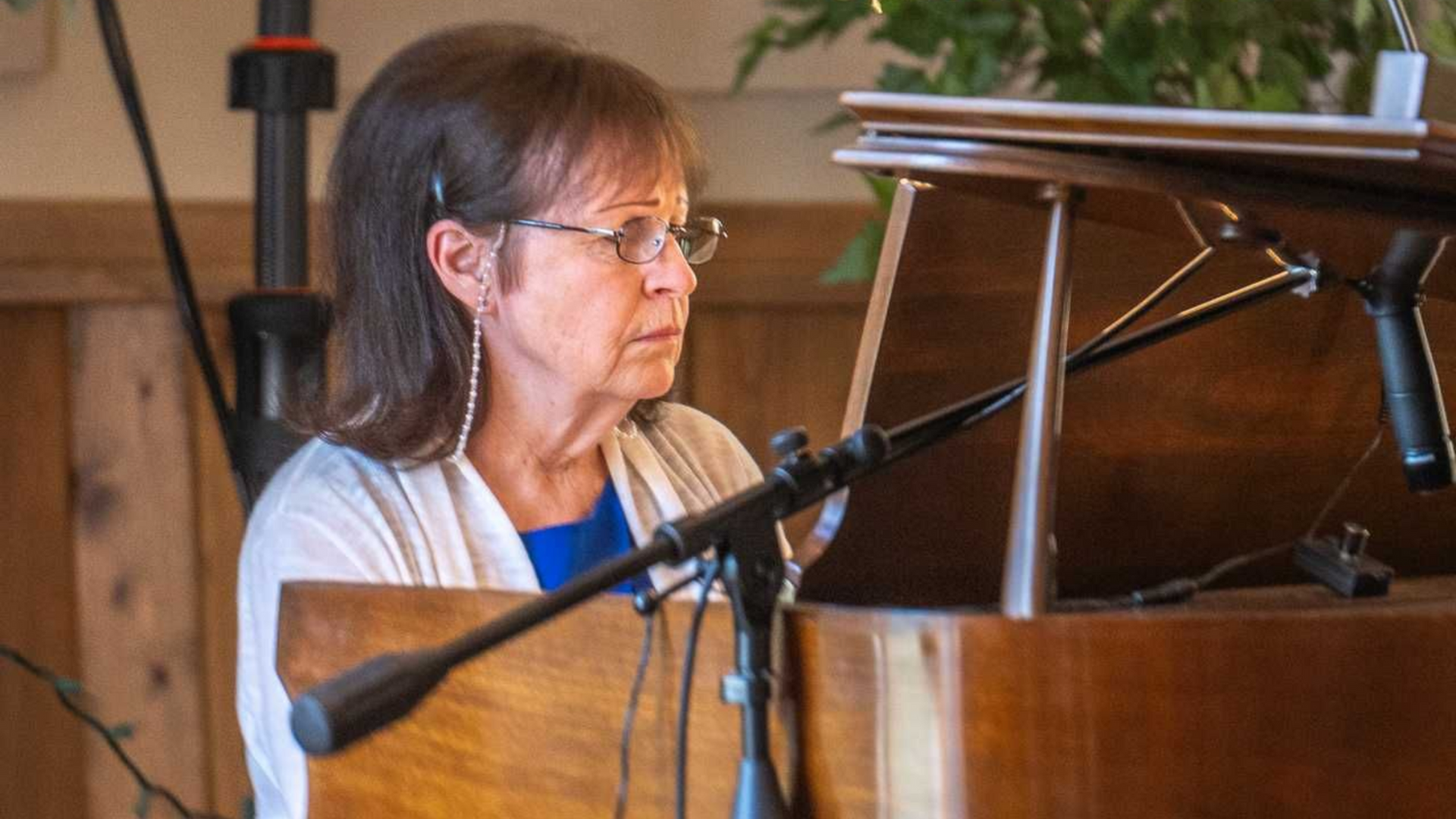Woman playing piano, wearing glasses, blue shirt, and cardigan. Microphones in front of instrument.