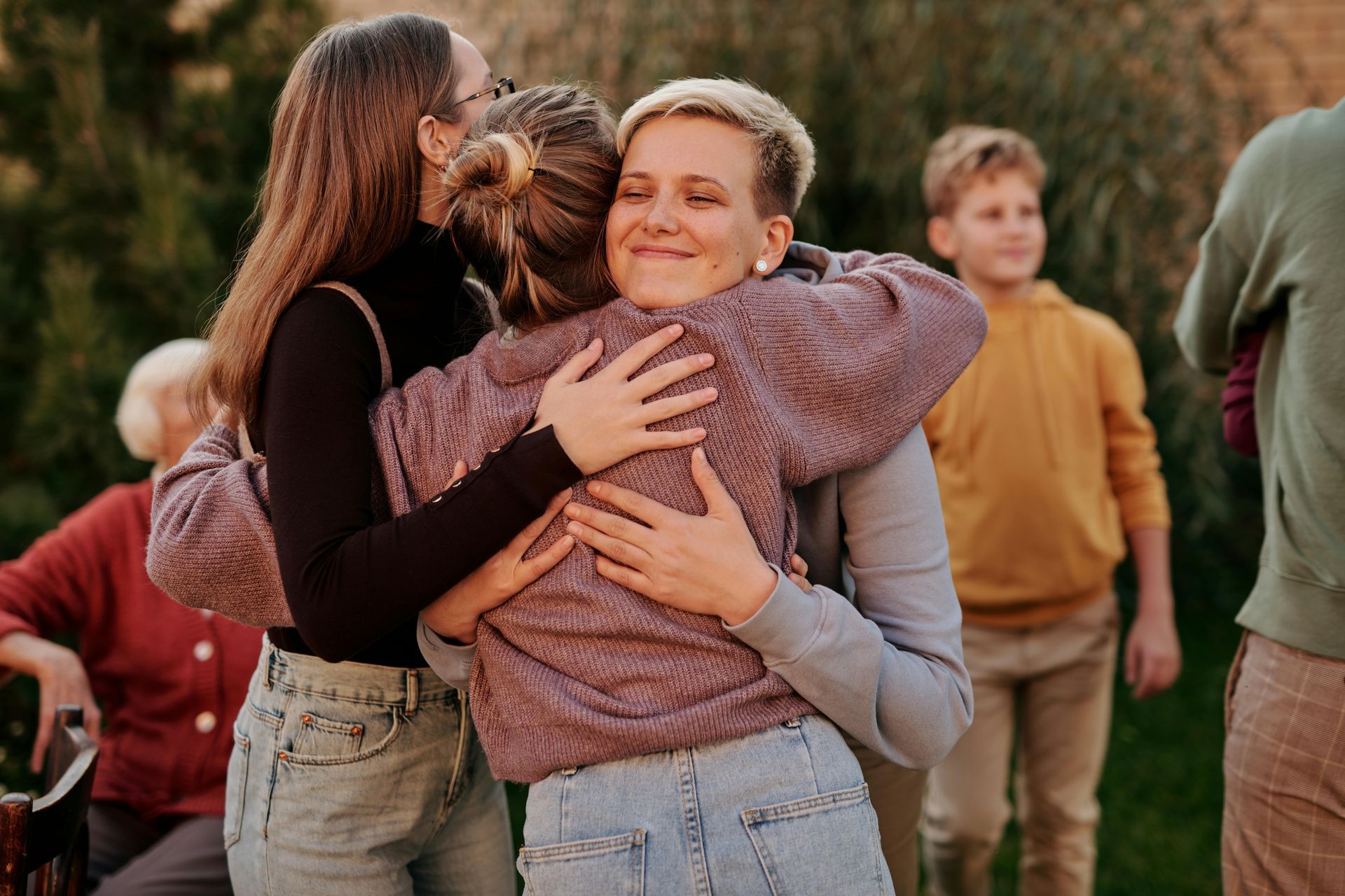 Two women embrace in an outdoor setting, smiling. Other people watch, golden hour light.