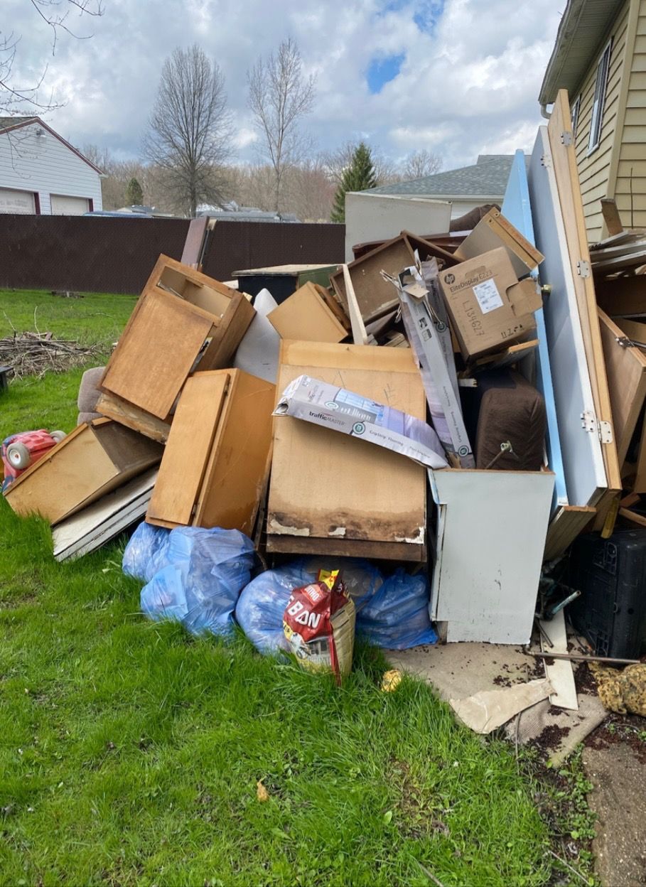 Piled cardboard boxes and trash bags beside a house on a grassy yard under a cloudy sky