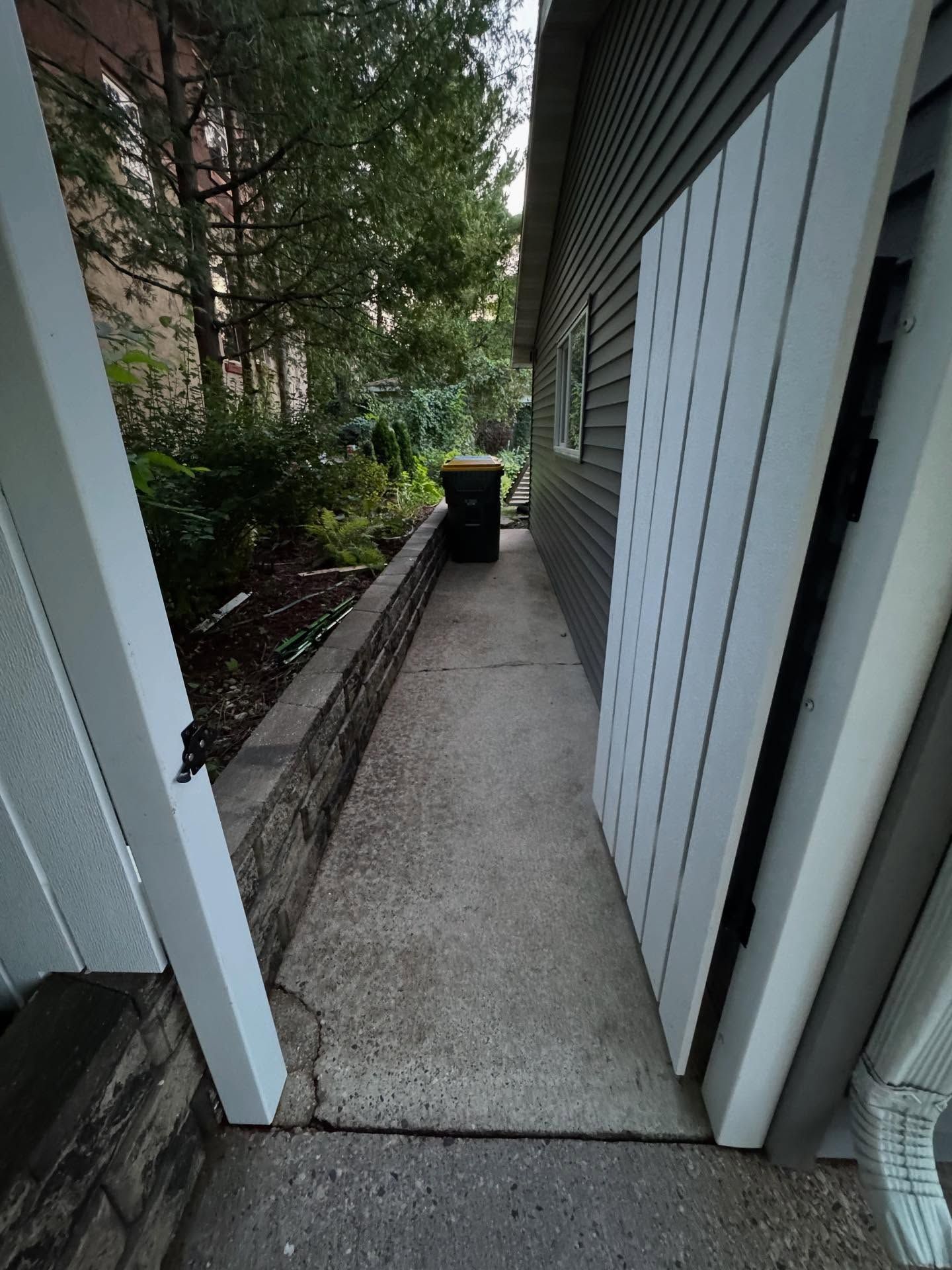 Narrow concrete side walkway beside a house, with a brick wall, white siding, and a trash bin ahead.