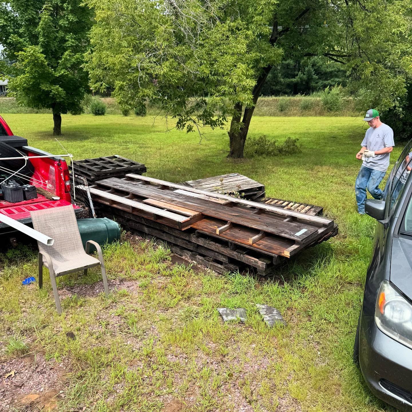 Man in blue shirt beside stacked wooden beams in a grassy yard, with trucks and trees nearby