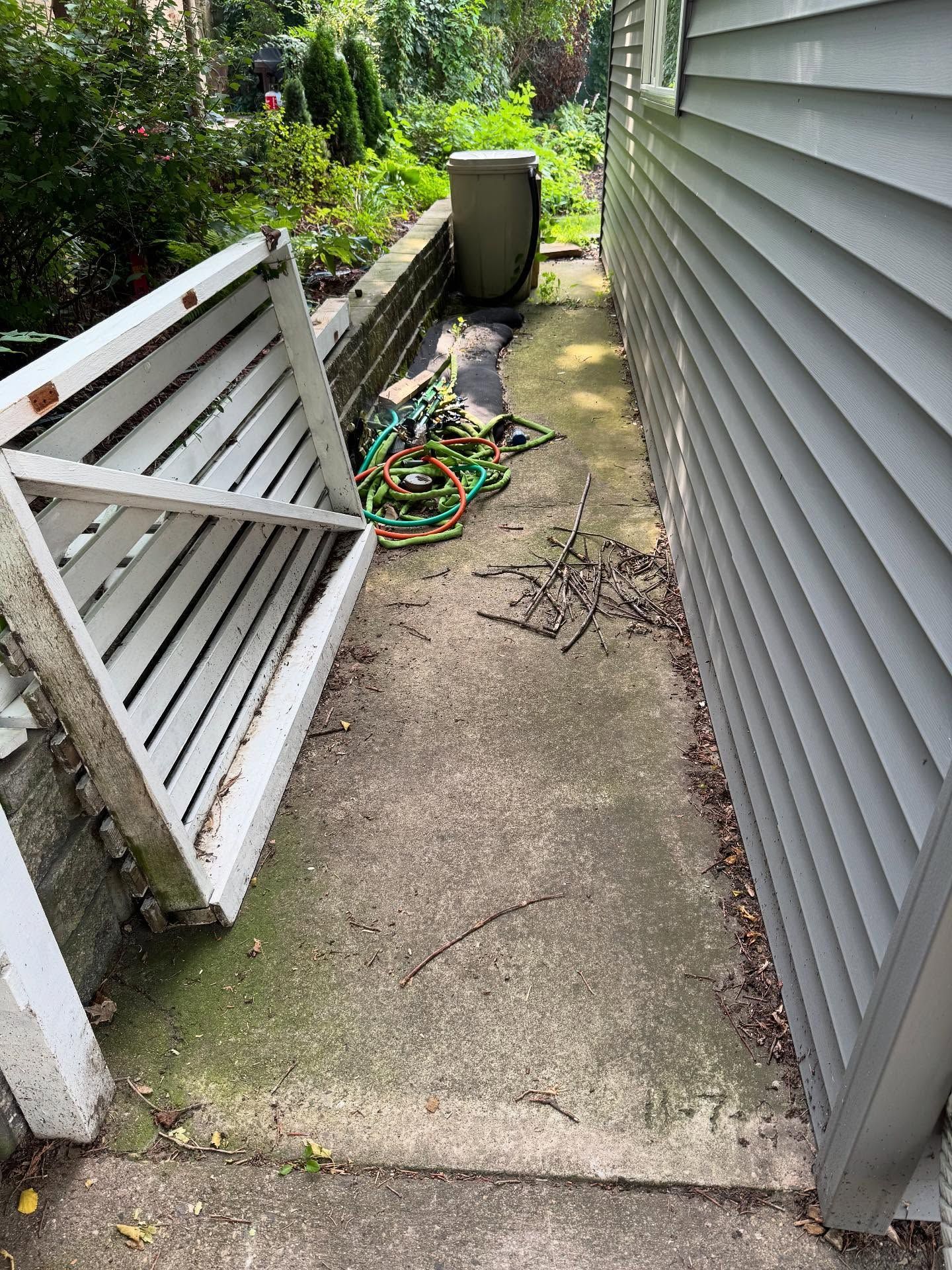 Narrow side yard with a leaning white fence panel, concrete path, garden hose, and trash bin beside a house.