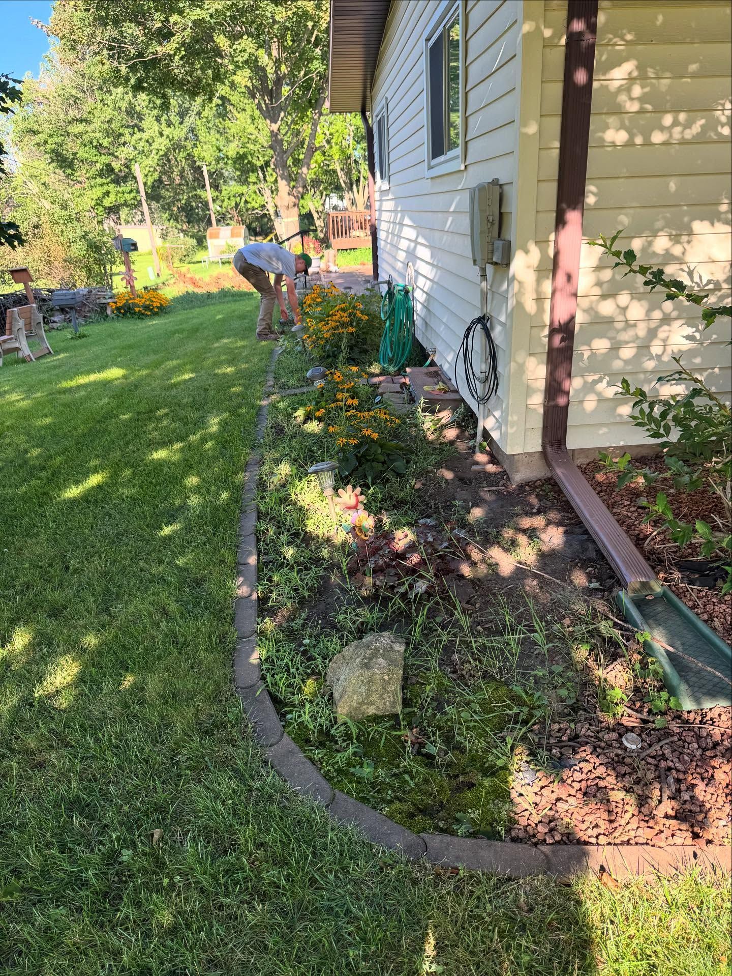 Person gardening beside a white house, tending a flower bed along a grassy yard