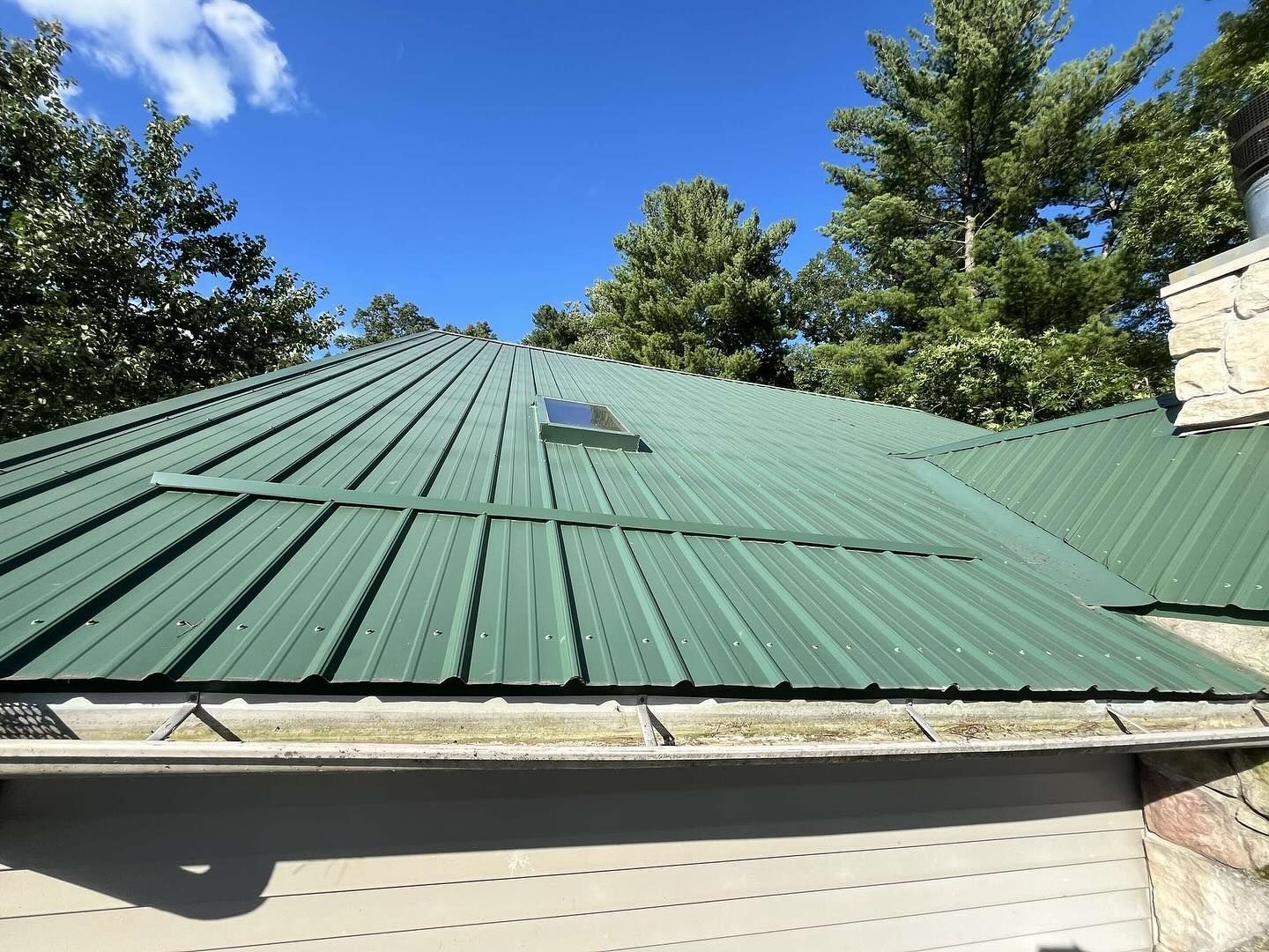 Green metal roof with a skylight under a bright blue sky, framed by trees