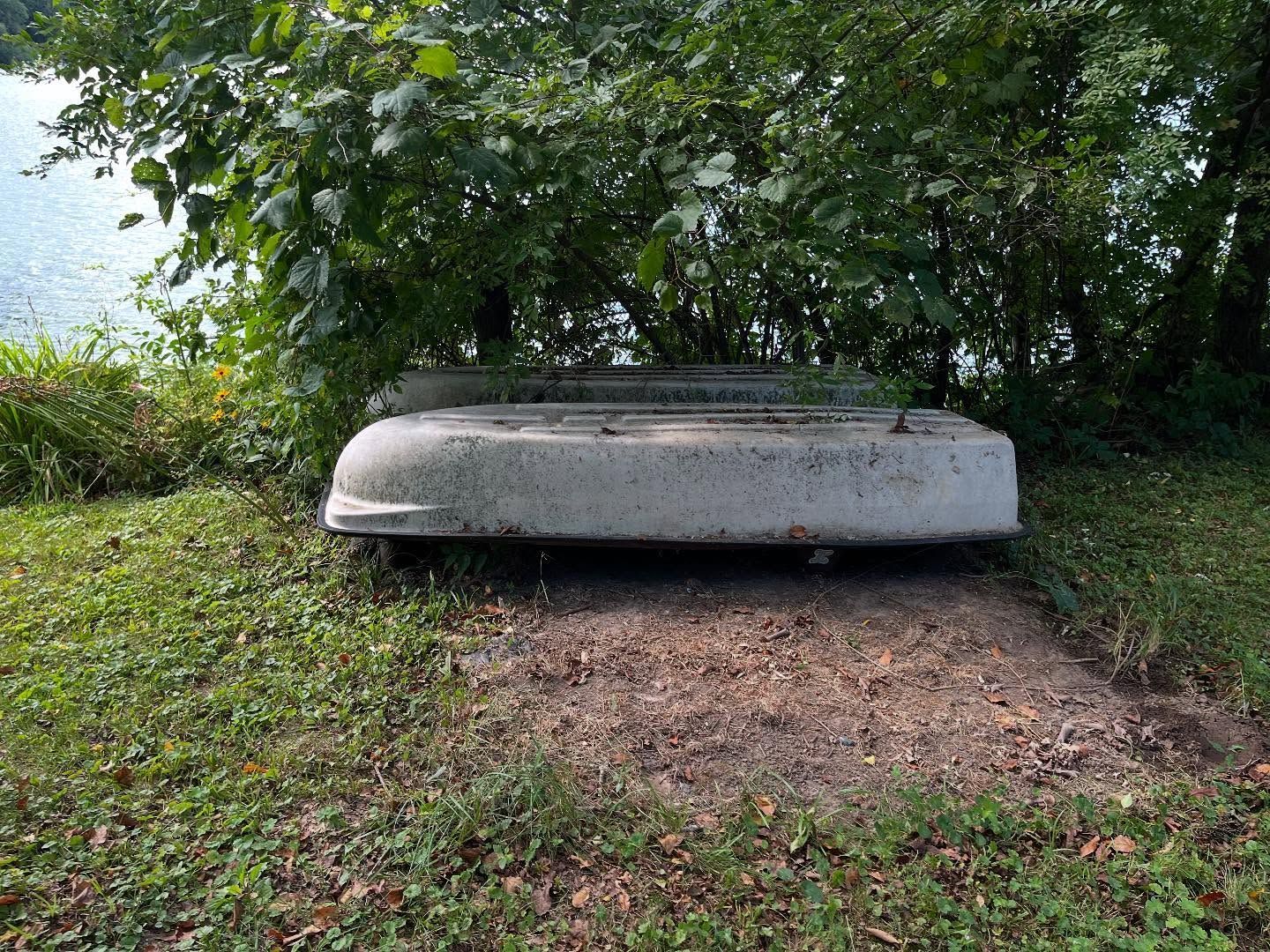 Overturned white boat hull in a grassy, wooded area by a dirt path
