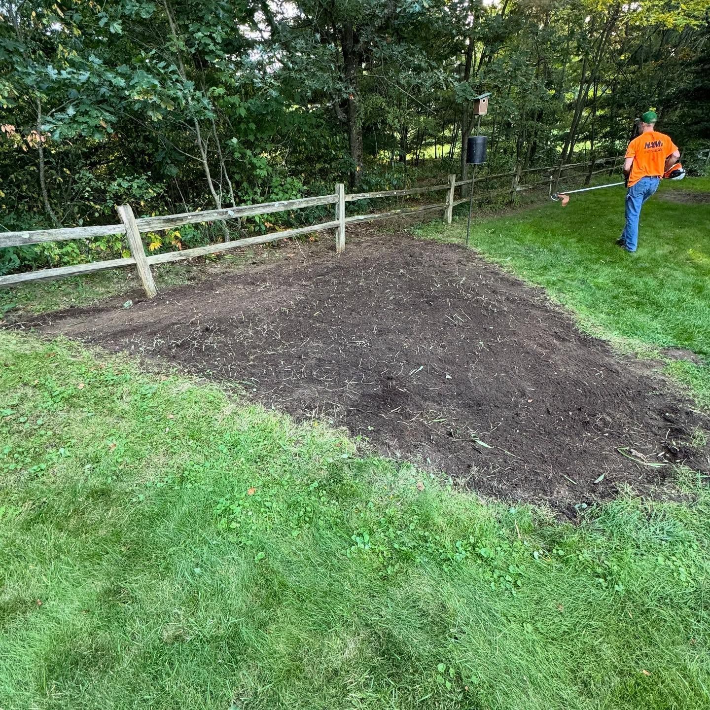 Freshly spread soil patch beside a white fence in a grassy yard, with a worker in an orange shirt nearby.