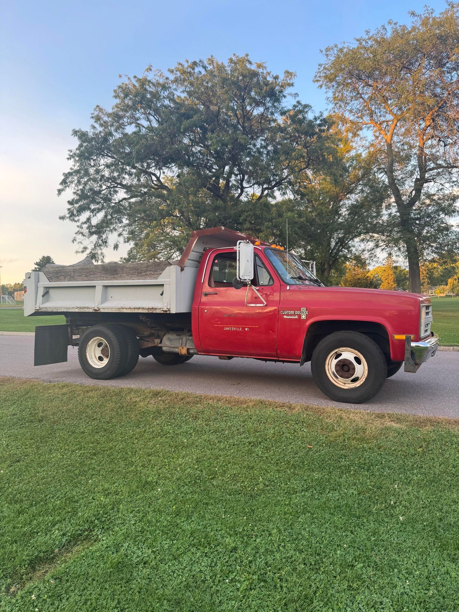 Red dump truck parked on a gravel driveway beside a grassy lawn at sunset.