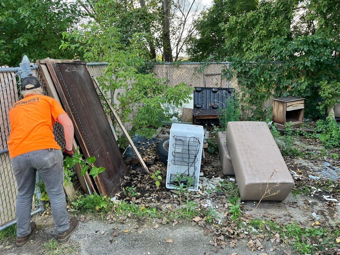 Person in orange shirt moving debris in a cluttered yard with stacked mattresses and overgrown greenery.