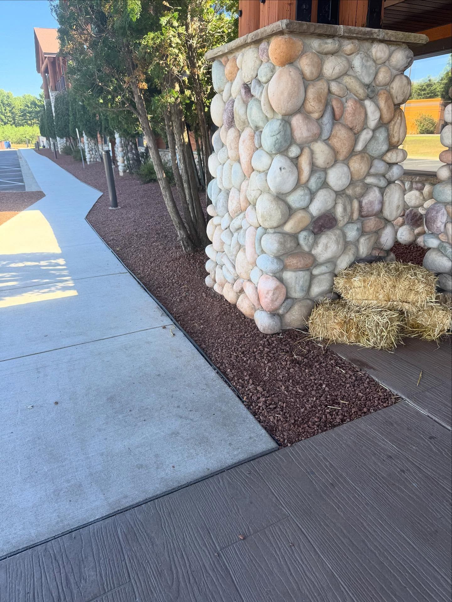 Stone pillar beside a sidewalk with landscaping and hay bales in a sunny outdoor area