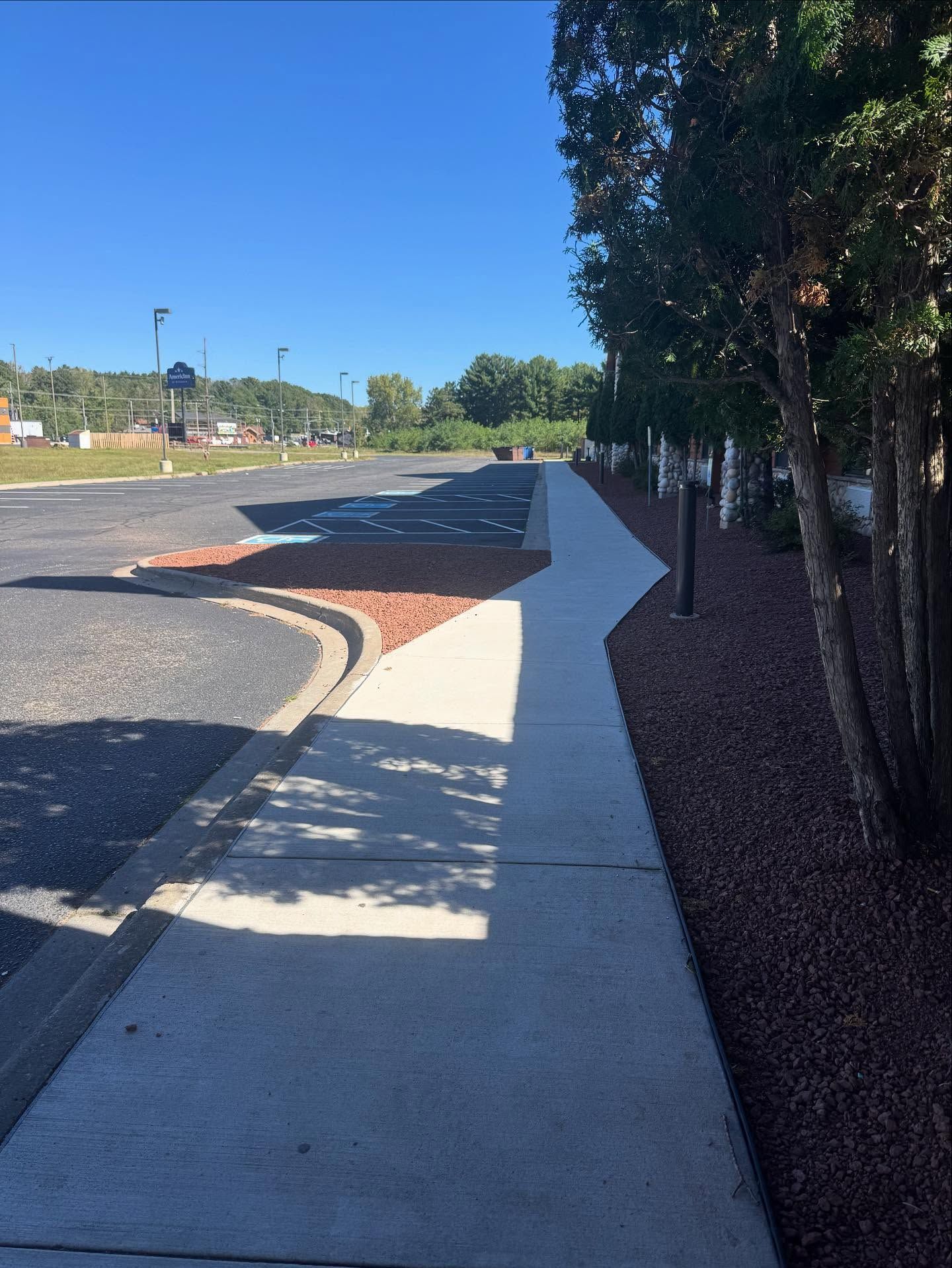 Empty parking lot beside a white sidewalk and dark fence under a clear blue sky.