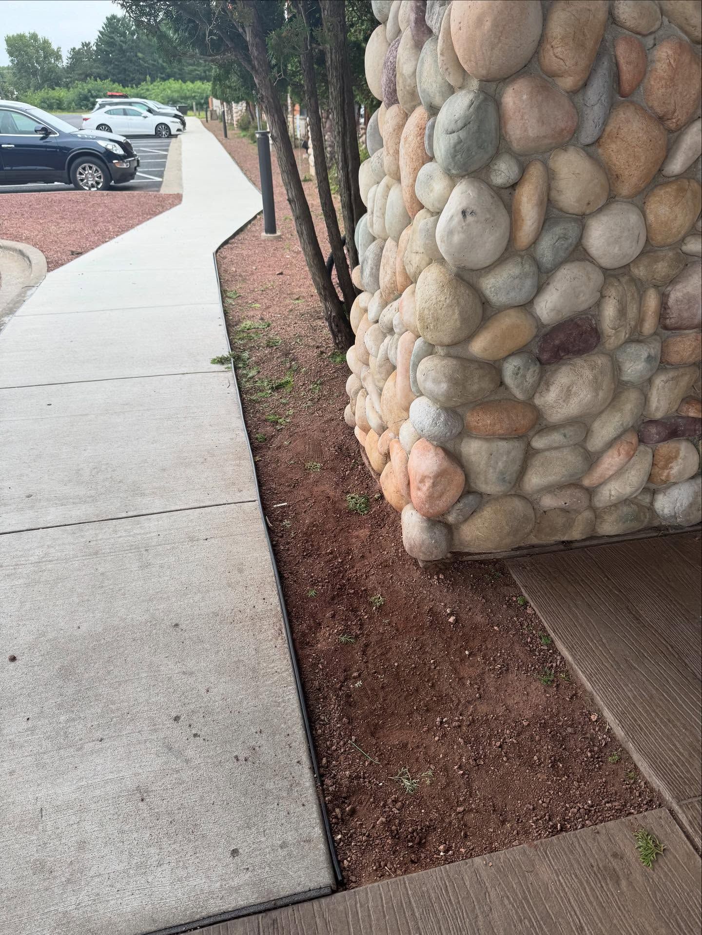 Sidewalk beside a stone column with a gravel strip, curving past parked cars and trees.