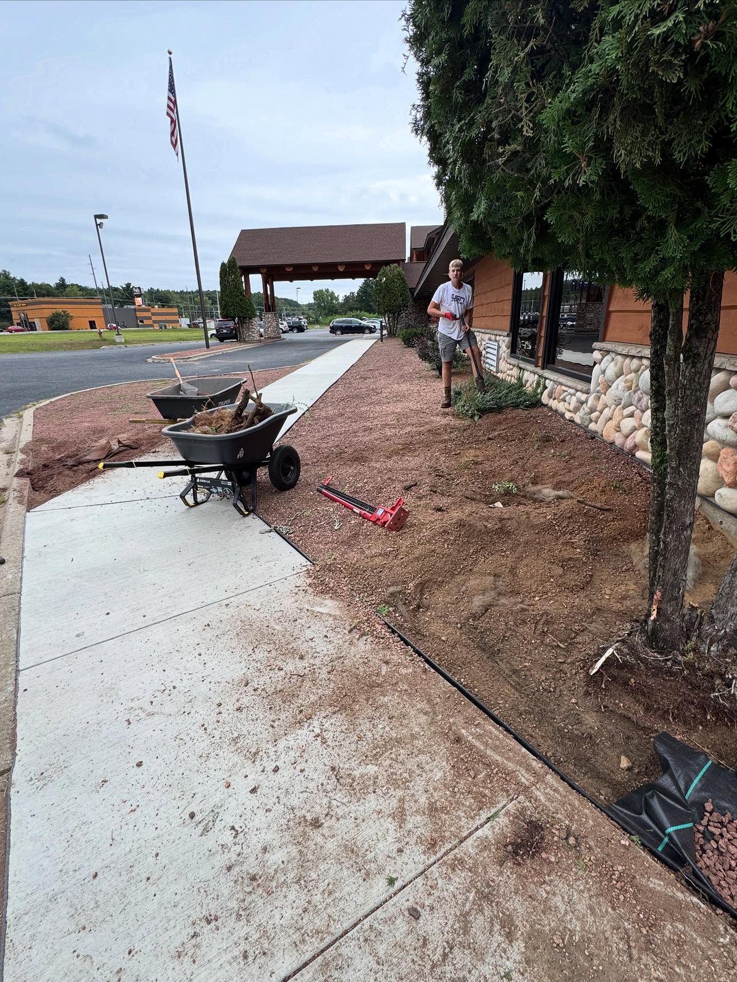 Damaged sidewalk beside a landscaped area with a wheelbarrow, dirt, and a worker near a covered entrance