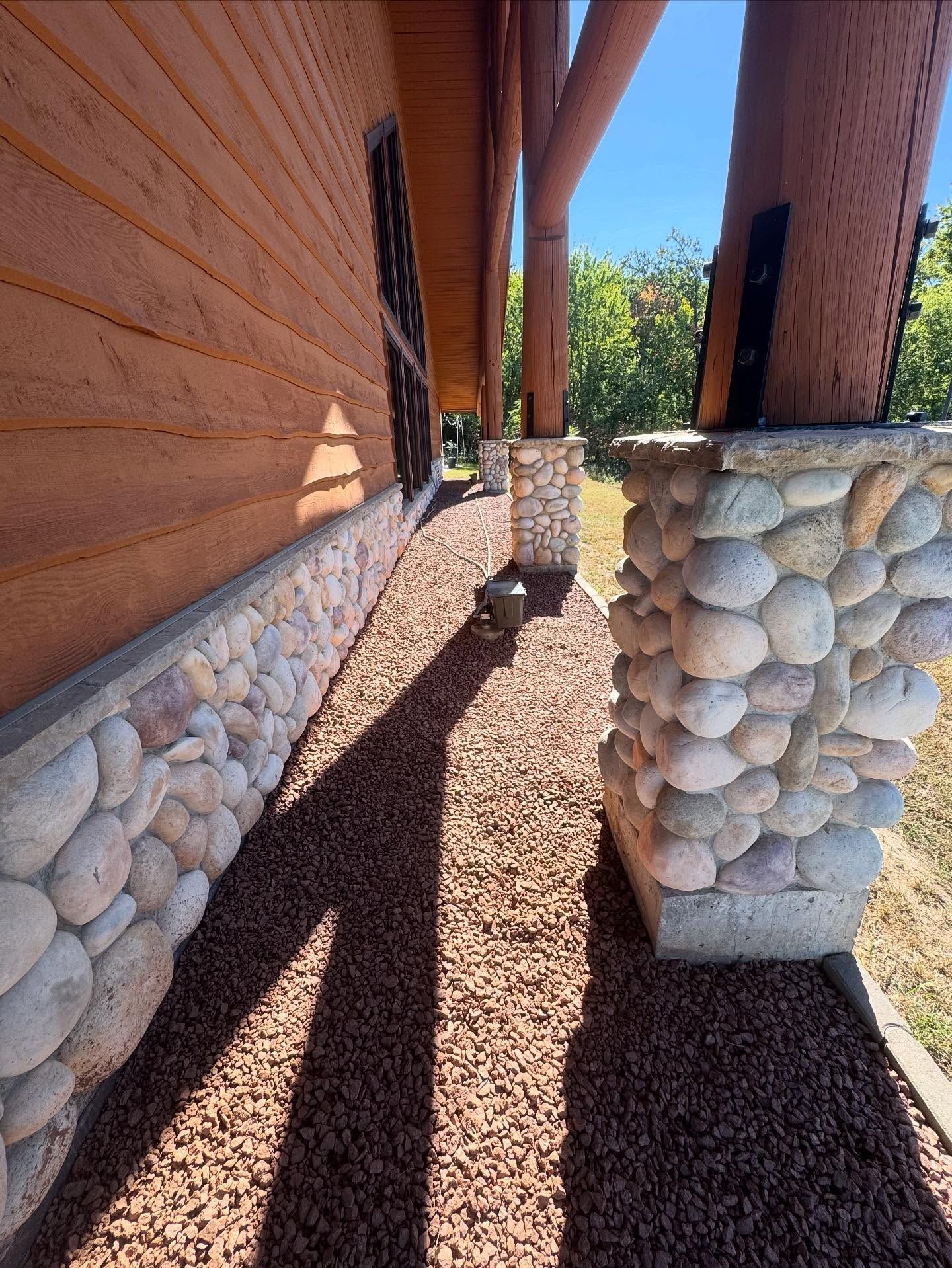 Narrow gravel path between stone and wood-clad walls under tall wooden posts in bright sunlight