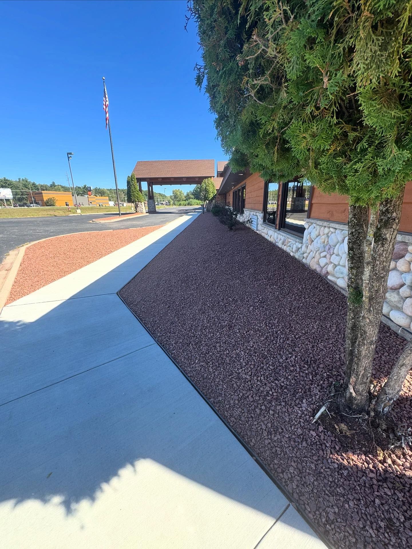 Sidewalk beside a building with trees, gravel landscaping, and a covered entry under a clear blue sky