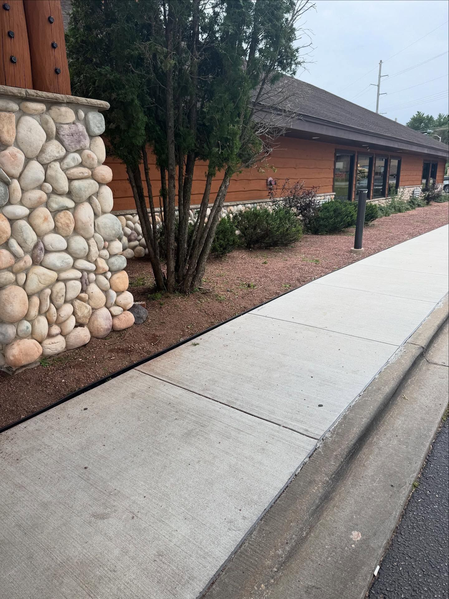 Sidewalk beside a brick building with stone column, shrubs, and a curb along the street.