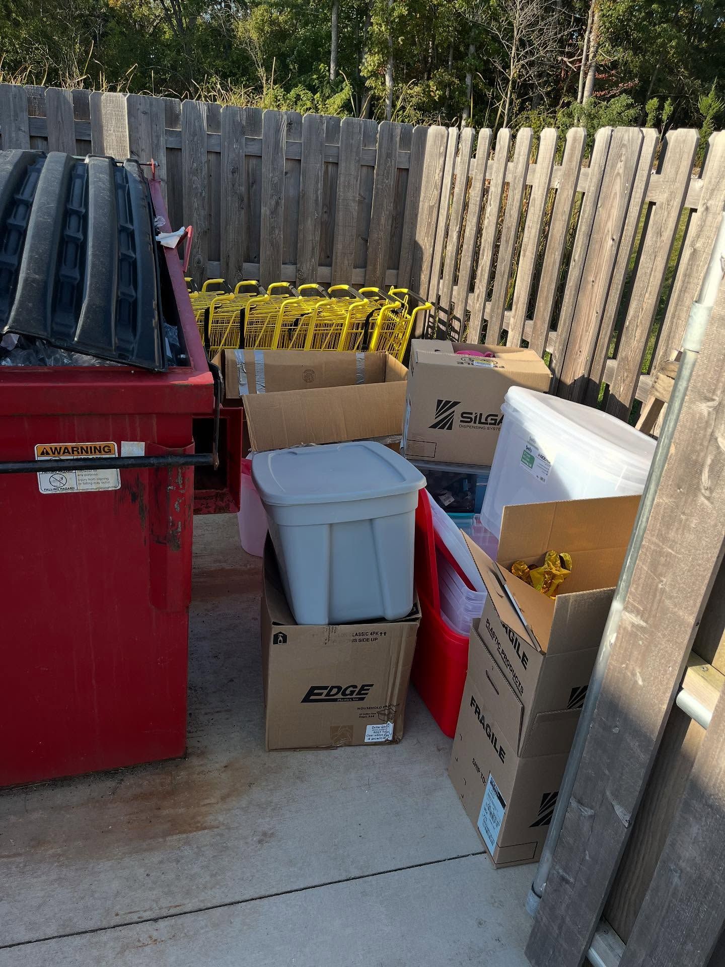 Outdoor fenced area with a red dumpster, stacked cardboard boxes, and white storage bins on a concrete patio.