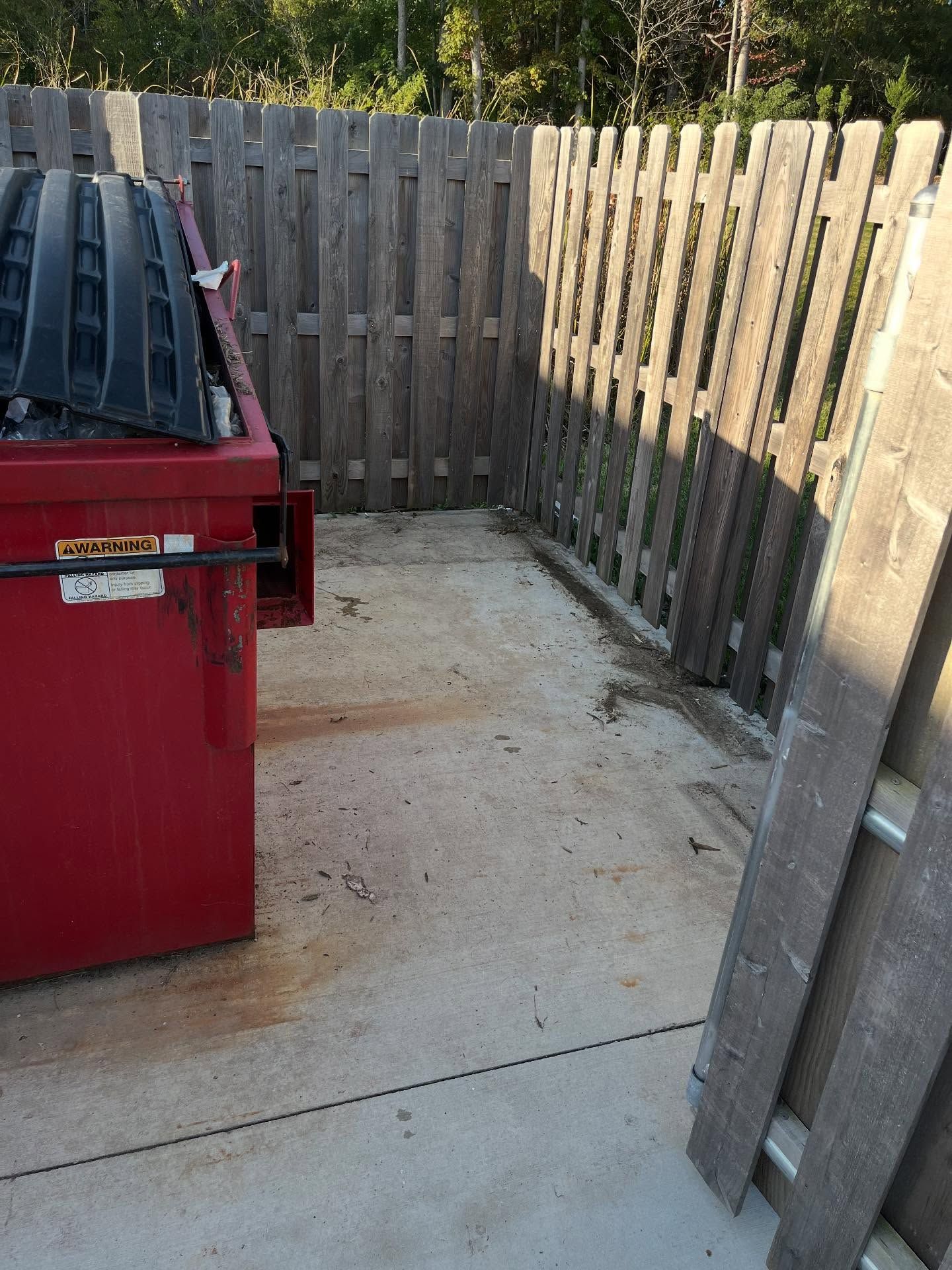 Concrete patio beside a wooden fence, with a red trash bin on the left and a leaning gate on the right.