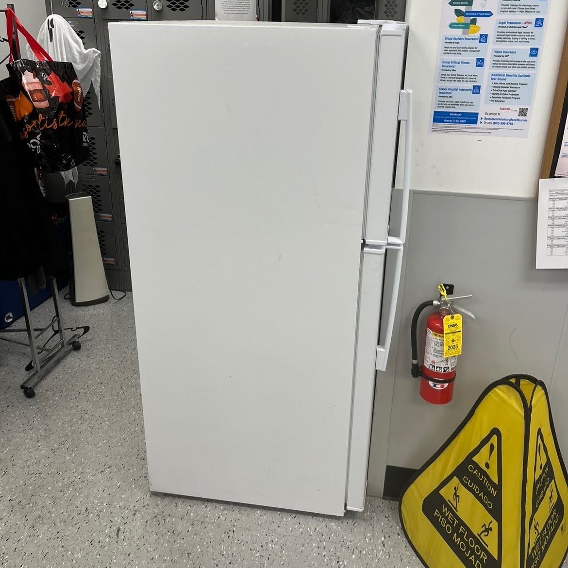 White refrigerator in a store aisle beside a fire extinguisher and wet floor sign.