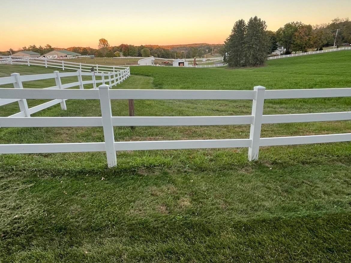 White wooden fence around a green pasture at sunset, with trees and a barn in the distance.