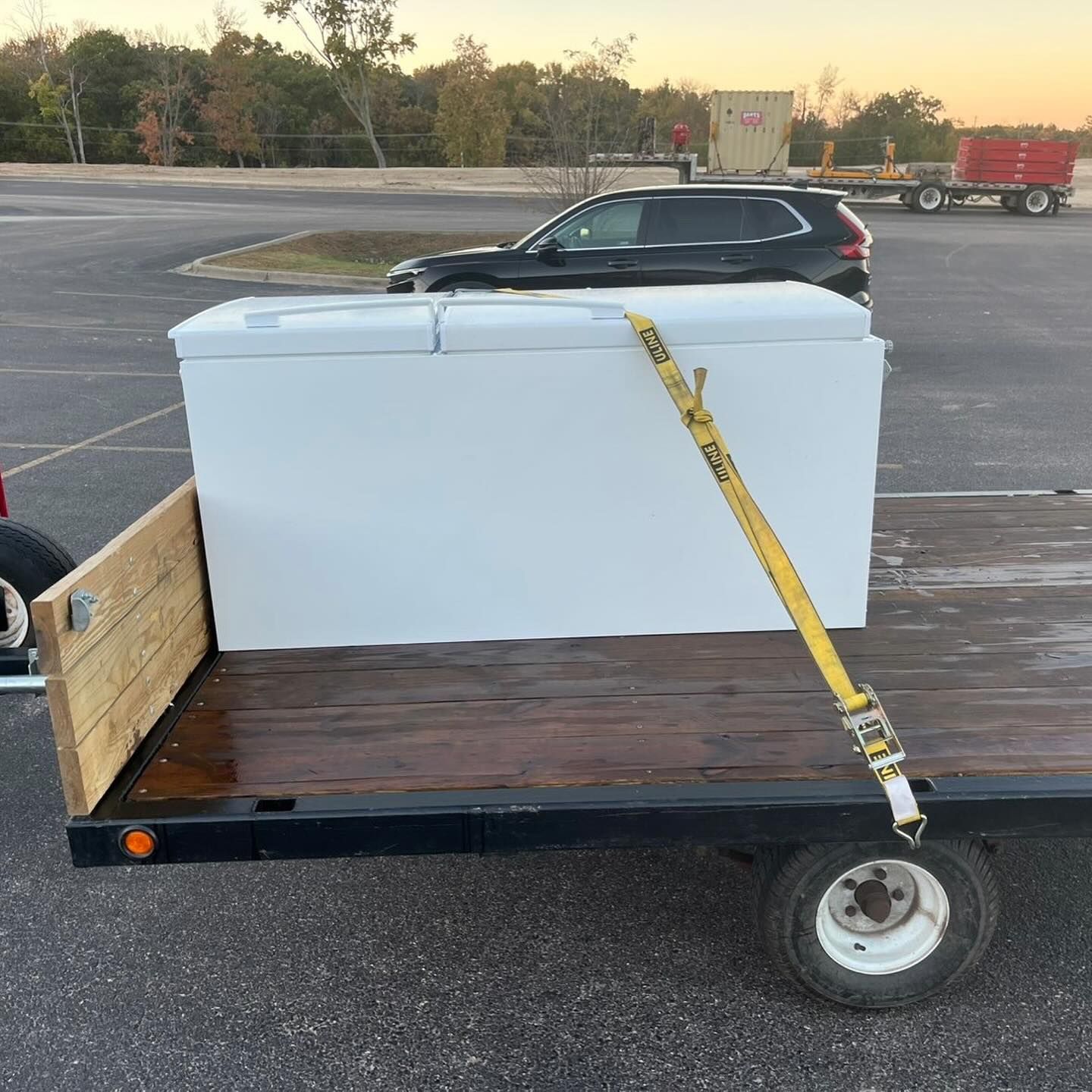Large white freezer strapped to a small utility trailer in a parking lot at dusk.