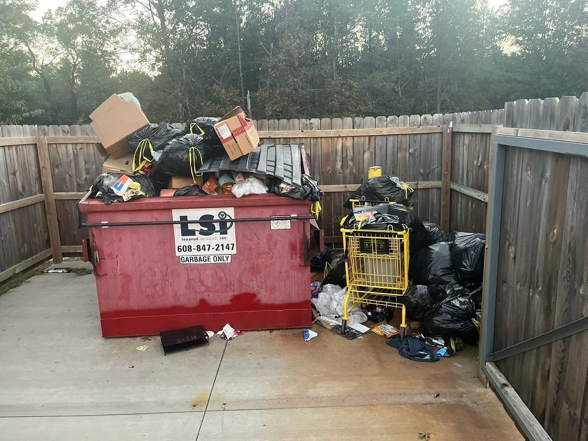 Overfilled red dumpster beside a wooden fence, with trash piled on top and scattered on the ground
