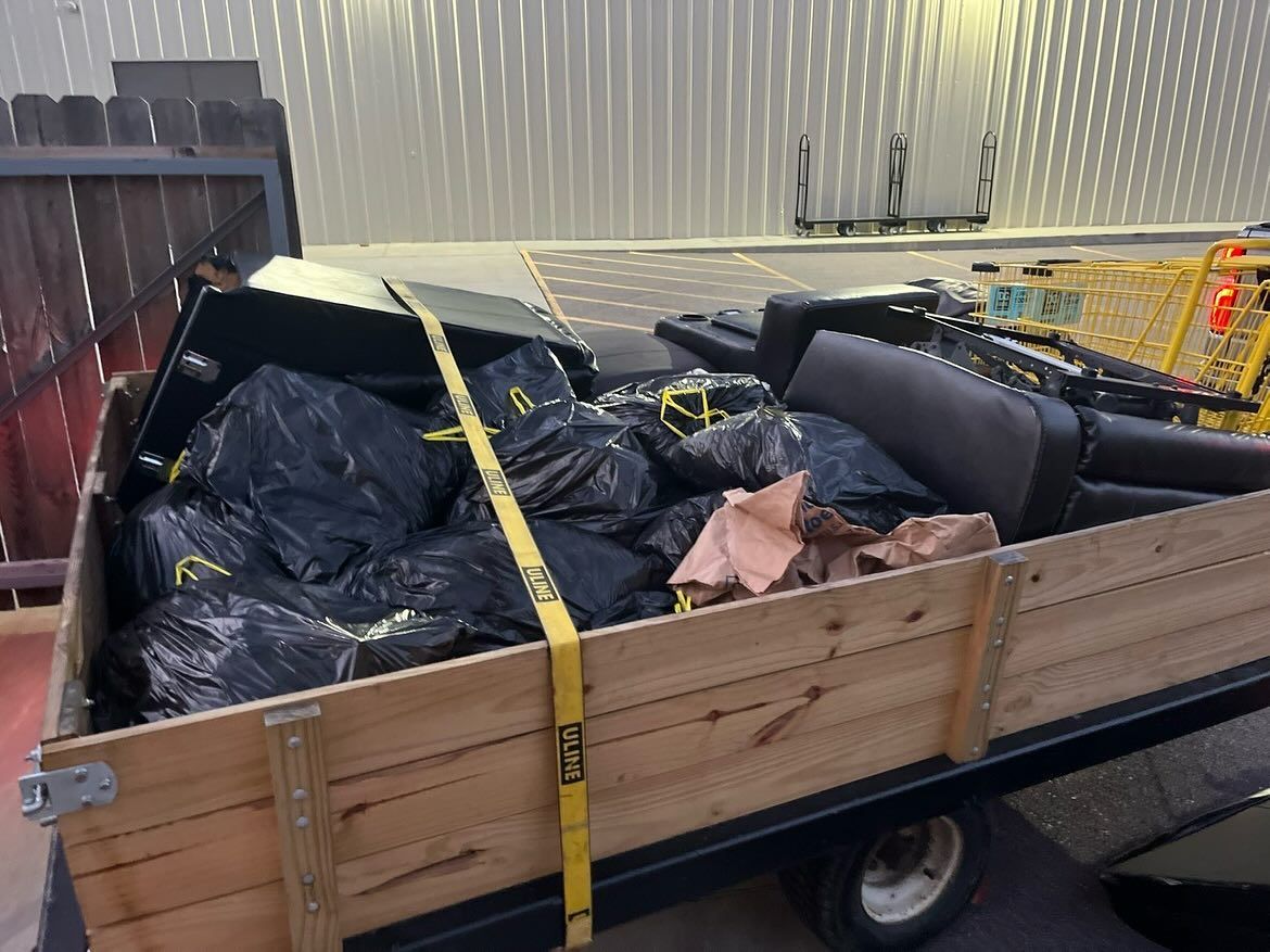 Wooden trailer loaded with black trash bags and debris in a driveway under a striped canopy