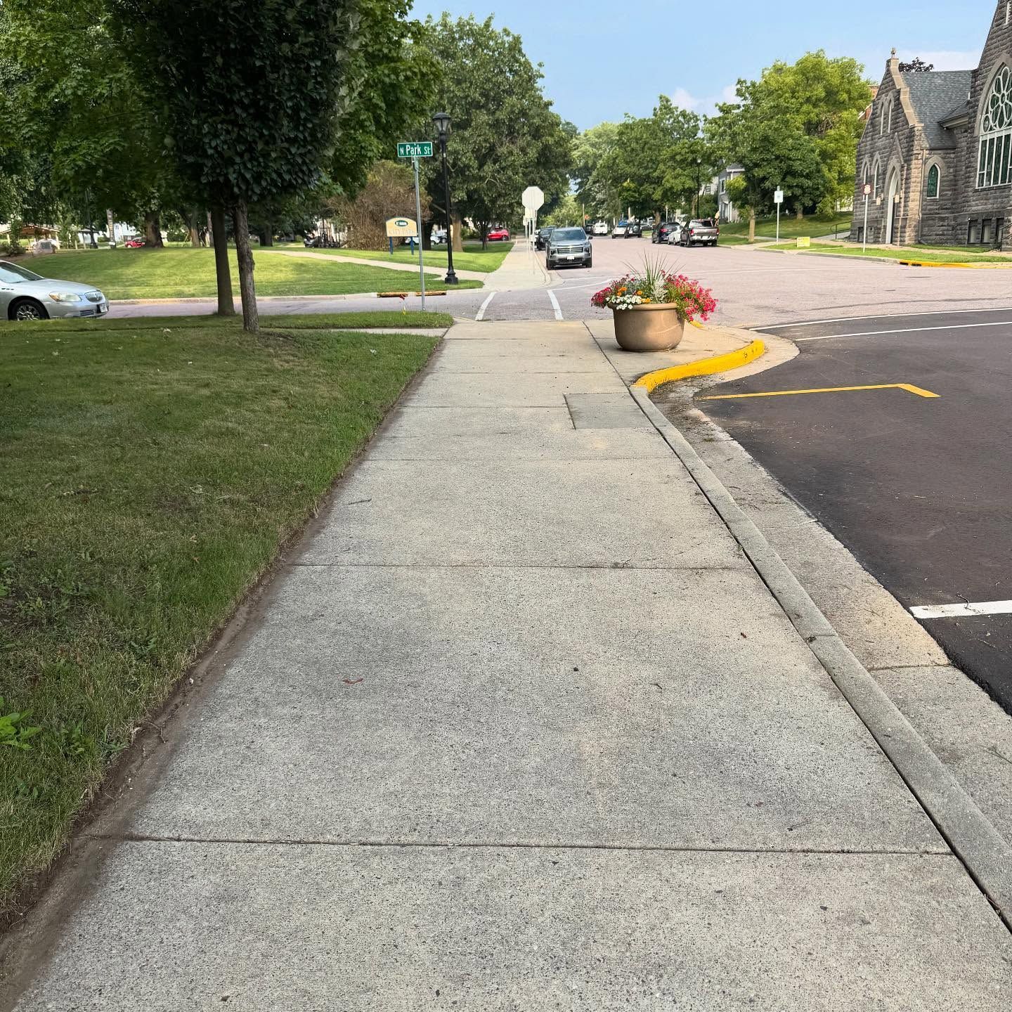 Sidewalk along a quiet street with grass, trees, parked cars, and a stone building on the right.