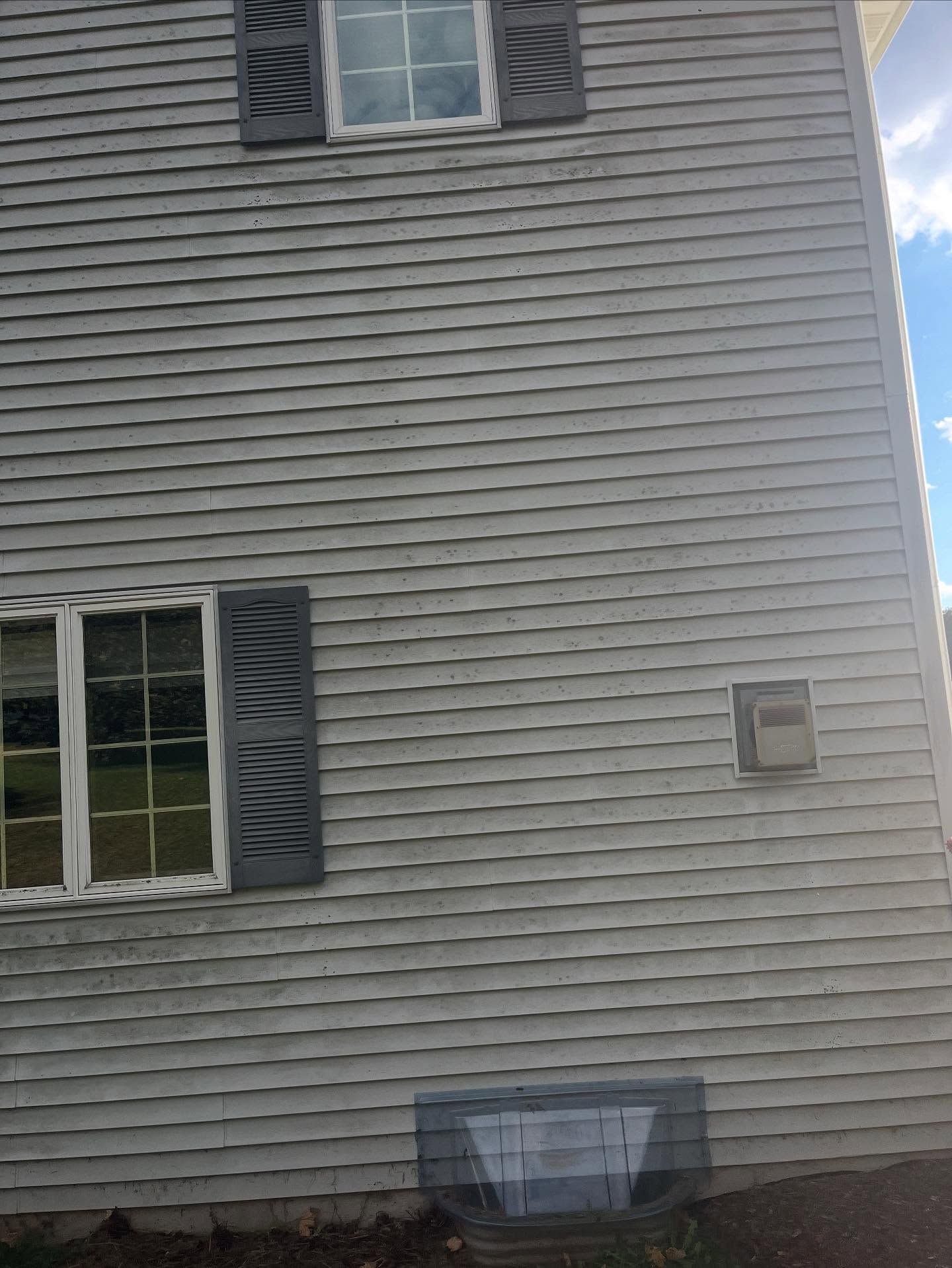 Side of a gray house with two windows, dark shutters, and a small basement window well.
