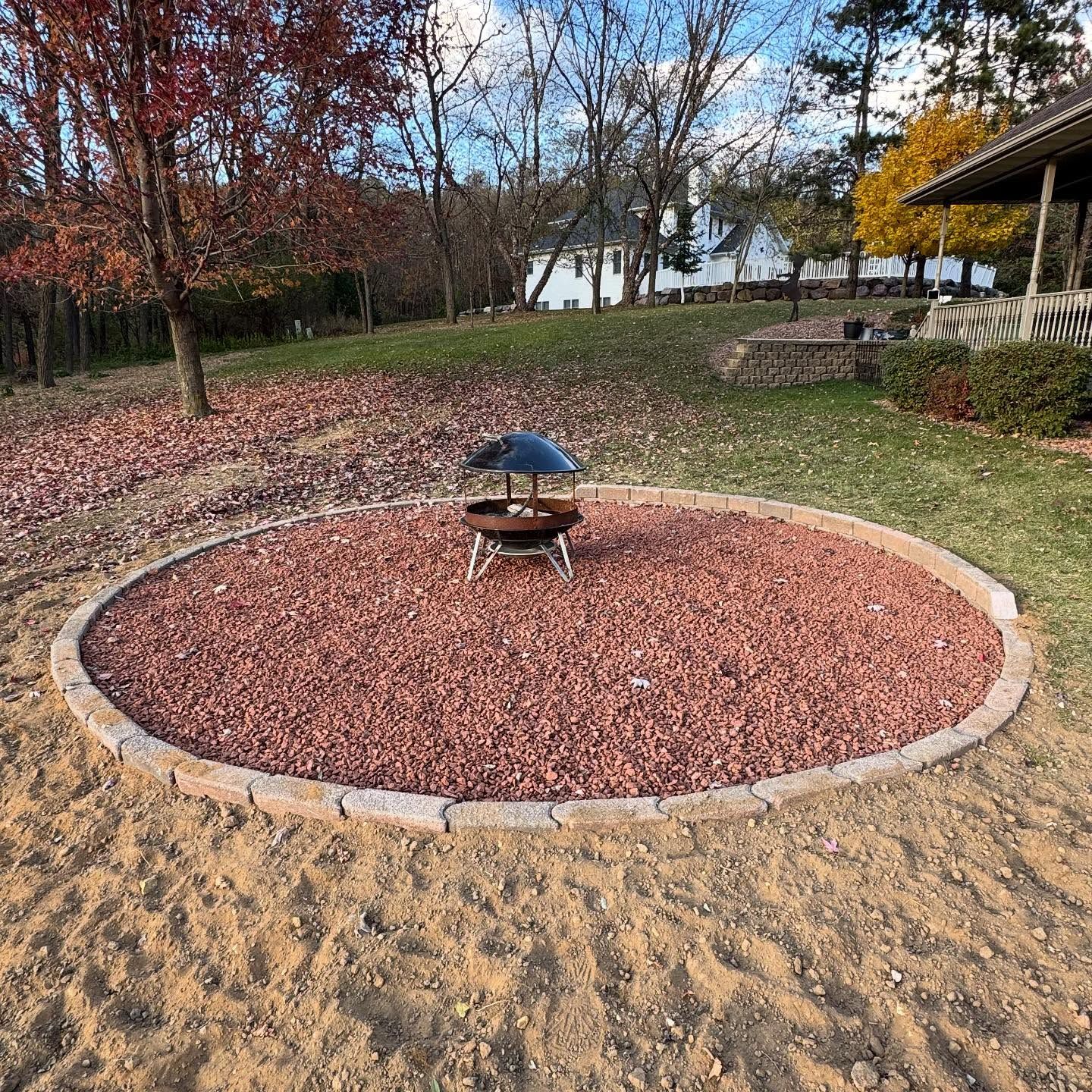 A circular gravel fire pit with a blue umbrella table in a wooded yard, bordered by sand and stone.