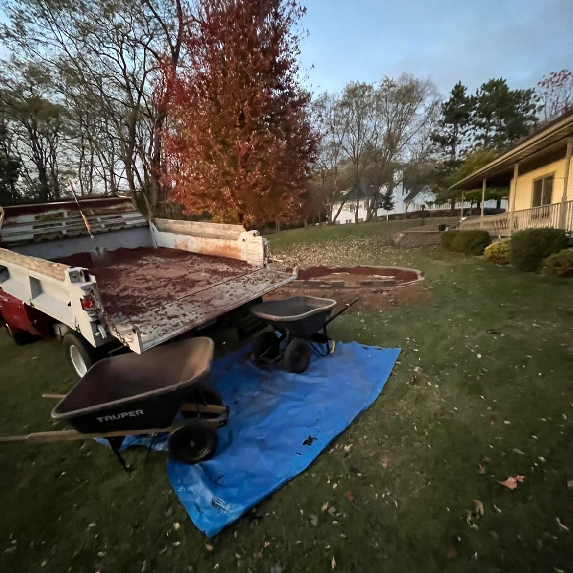 Black wheelbarrow and yard cart on a blue tarp beside a pickup truck in a grassy yard at dusk