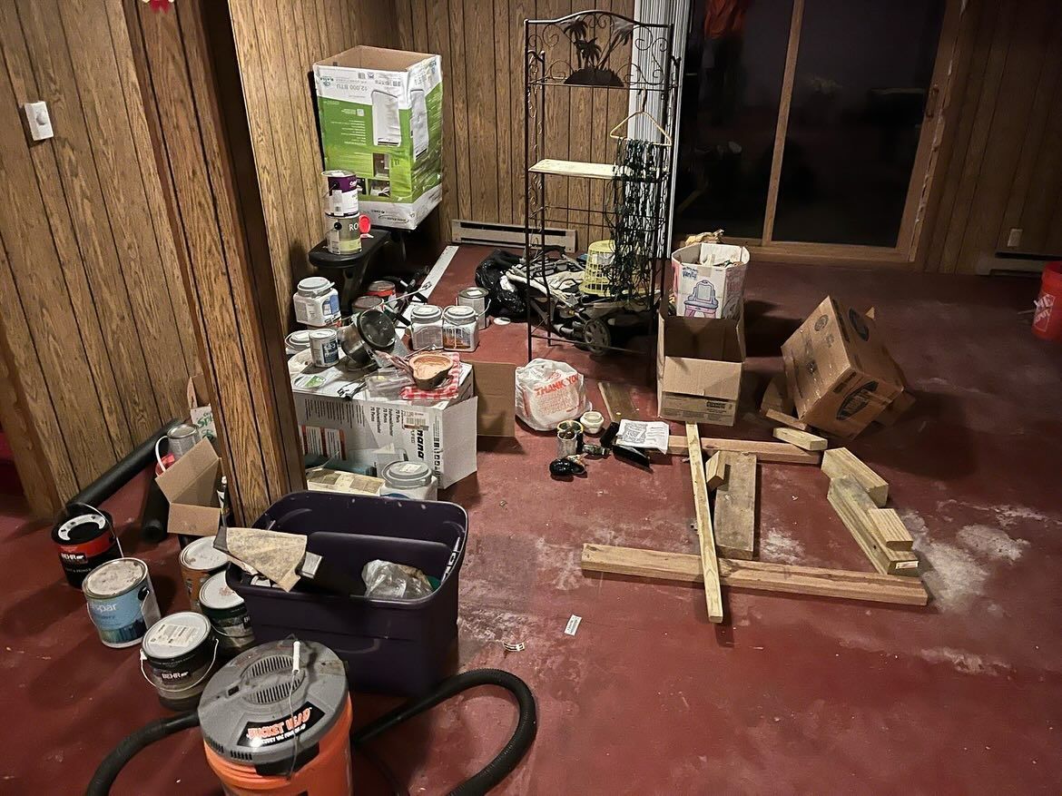 Cluttered workshop floor with tools, paint cans, boxes, and wooden materials near a doorway.