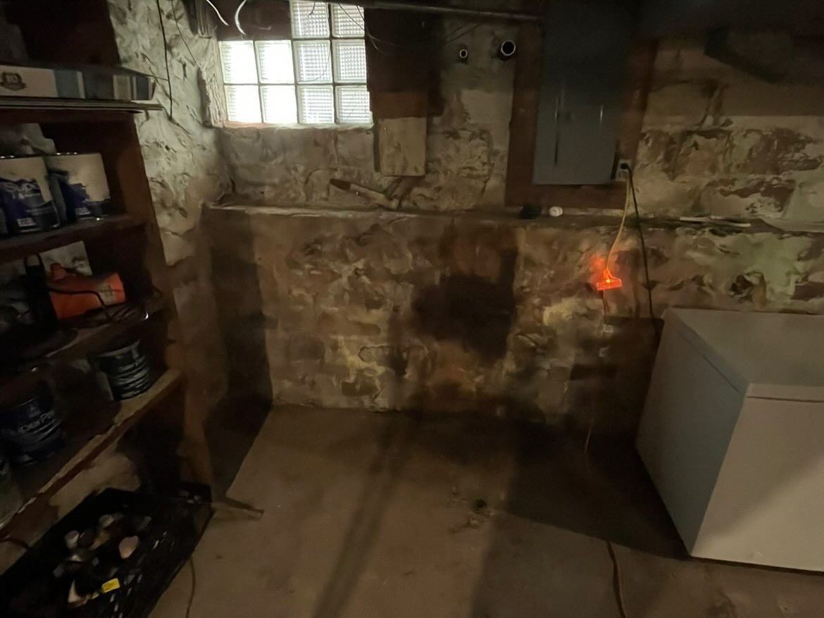 Dim unfinished basement with stained concrete walls, shelves on the left, and a washing machine on the right.
