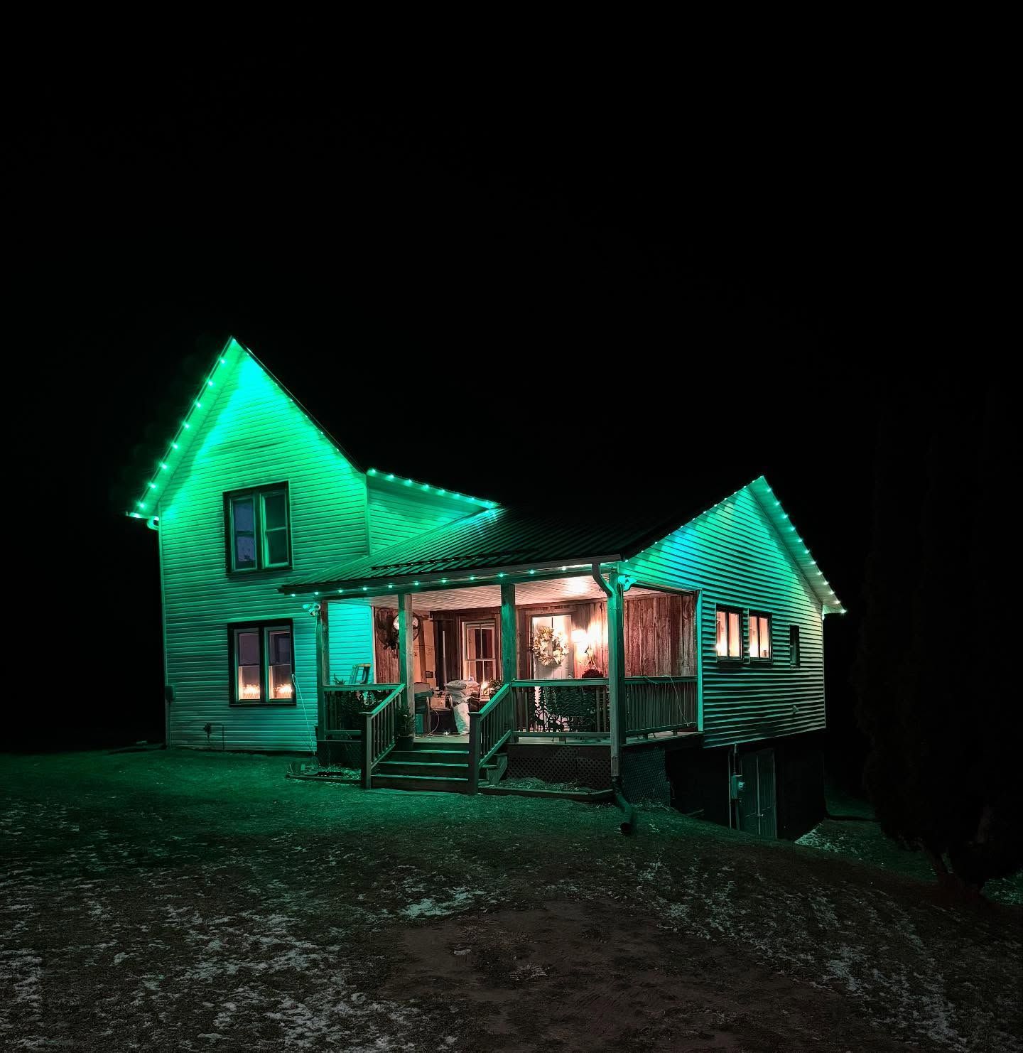 Small wooden house with a lit porch and green neon roof lights at night on a dark hillside.